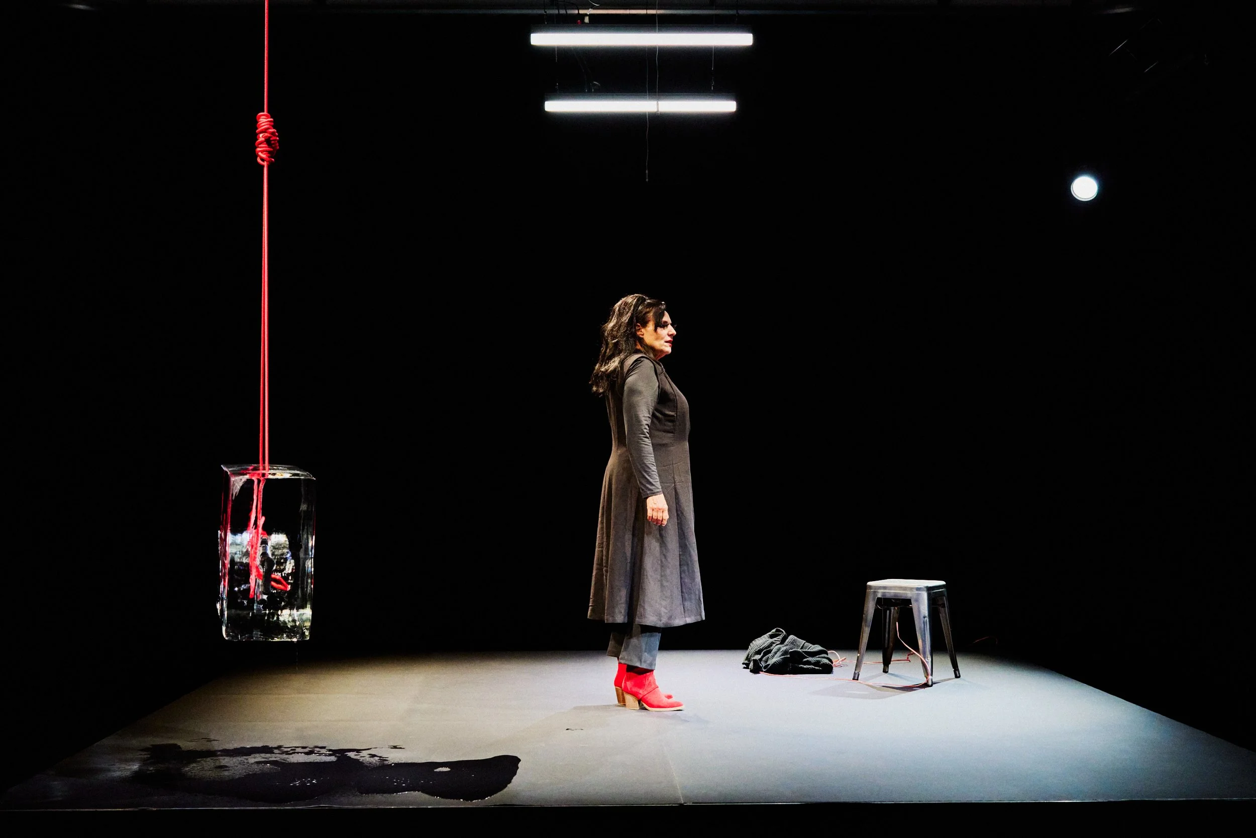 Eine Frau steht auf einer beleuchteten Bühne, umgeben von einem Hocker, einem schwarzen Kleidungsstück und einem schwarz-weißem Mann. Es gibt eine Skulptur mit roter Schnur und einem schwarzen Fleck auf dem Boden.