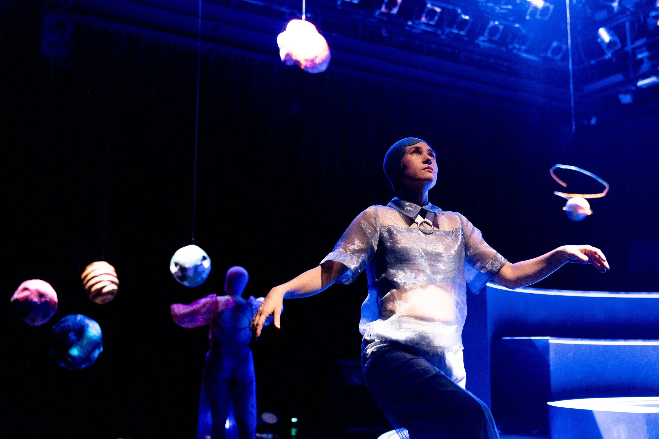 Eine Person auf einer Bühne, umgeben von schwebenden Kugeln und einem Puppenspiel, bei einer Theateraufführung, mit dunklem Hintergrund und blauen Lichteffekten.