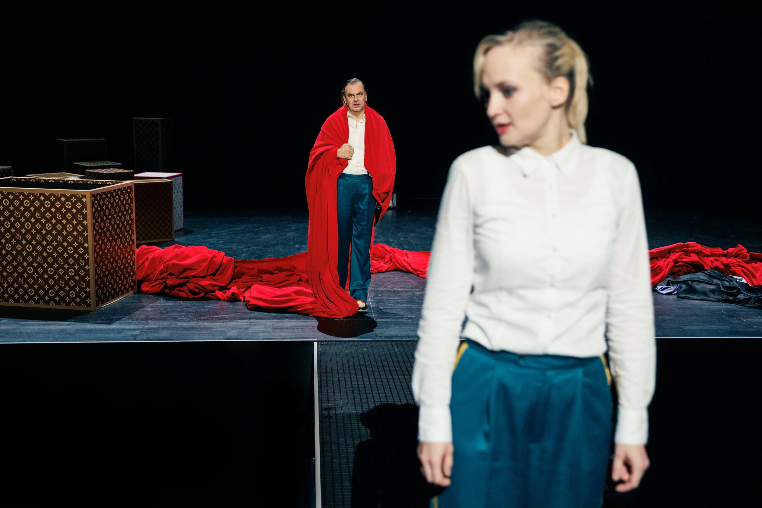 Zwei Schauspieler auf einer Theatbühne, einer im Hintergrund mit roten Stoff über die Schultern, der andere im Vordergrund, unscharf, in Weiß und Blau gekleidet.