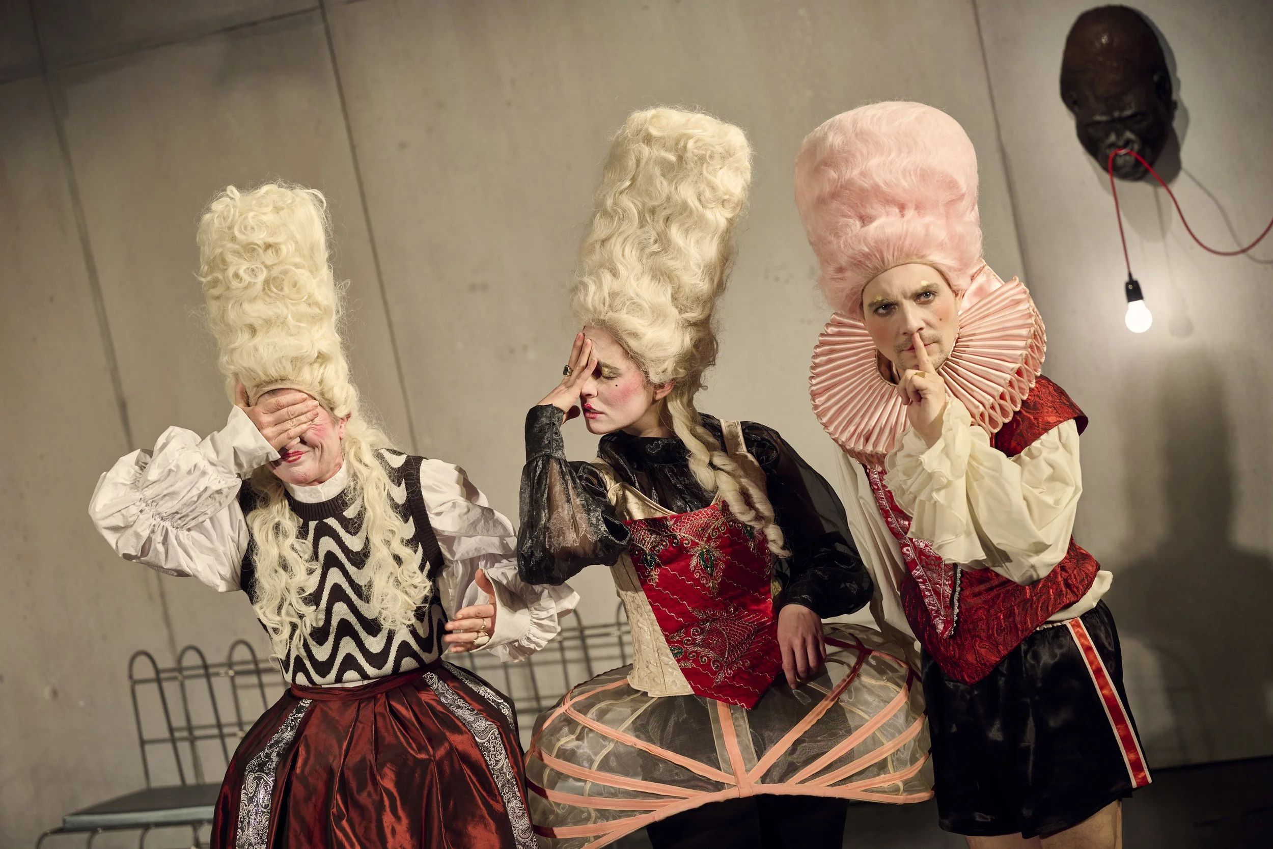 Drei Menschen in bizarrem Kostüm mit großen, ausgefallenen Perücken, die auf einer Bühne stehen und verschiedene Gesten machen.