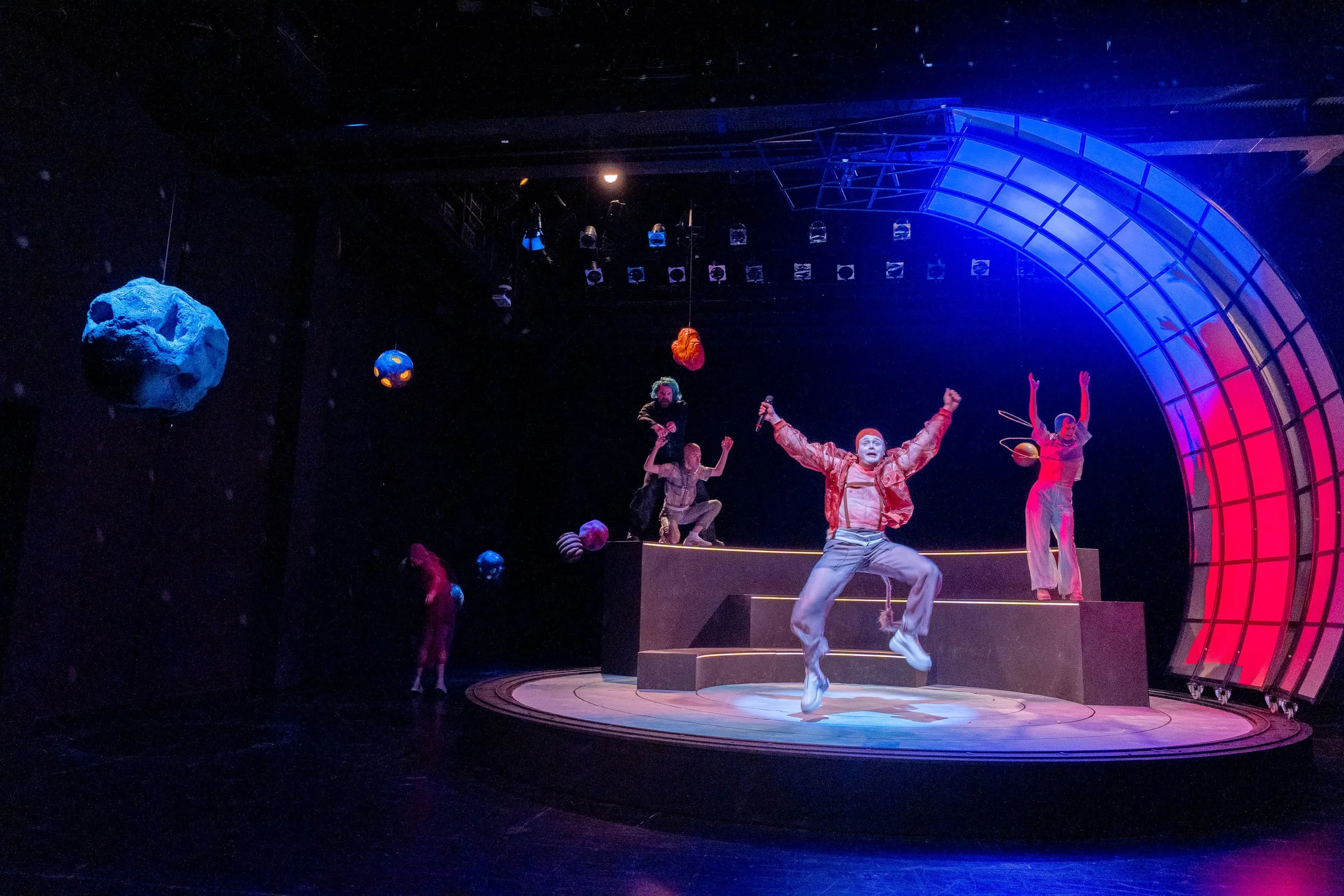 Bühnenszene mit mehreren Akrobaten und Schauspielern in bunten Kostümen, einige springen, während andere auf einem erhöhten schwarzen Pult stehen, im Hintergrund sind hängende Strukturen und dekorative Objekte, beleuchtet mit buntem Licht