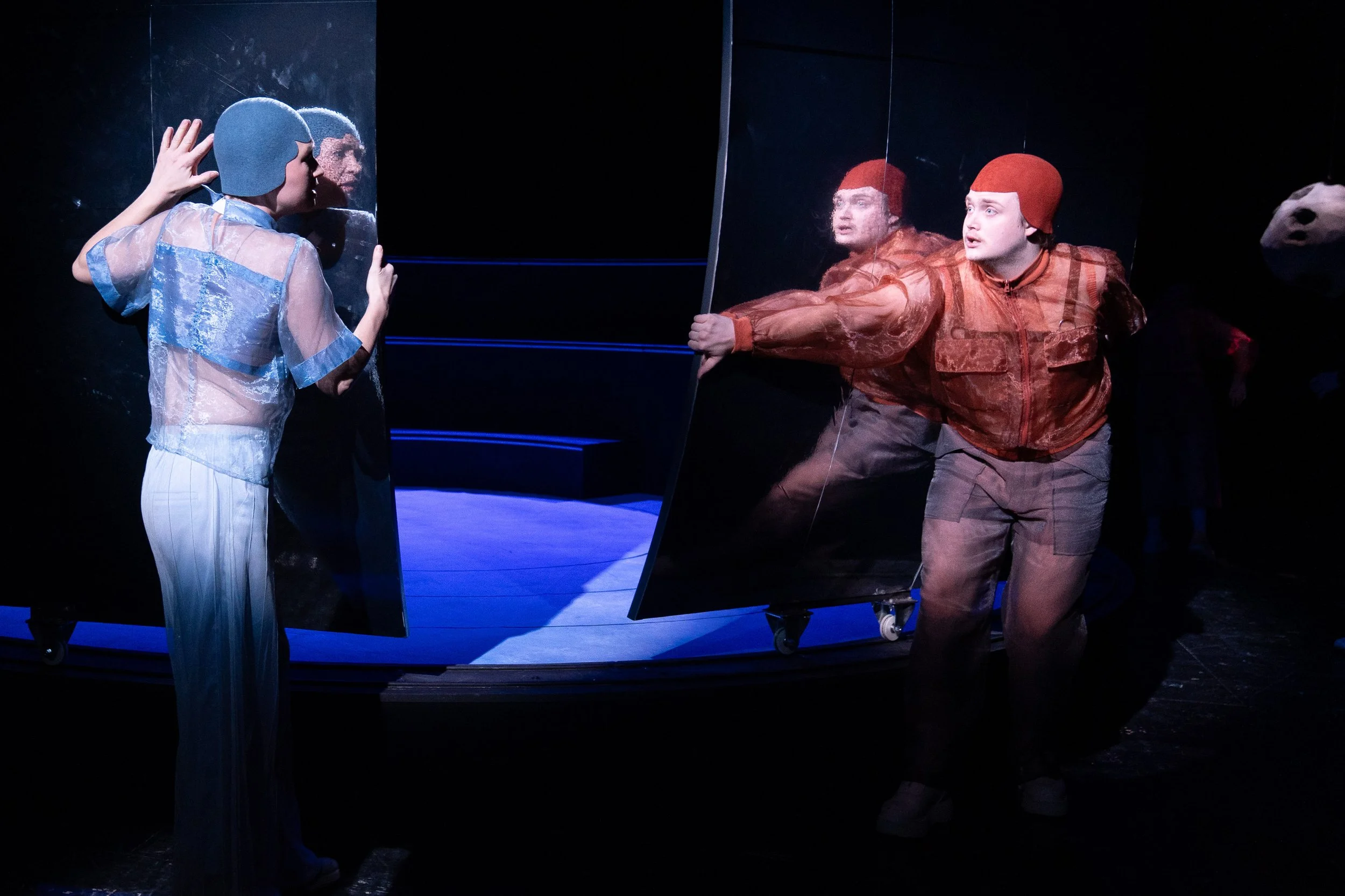 Zwei Schauspieler in Kostümen stehen vor einer tränenförmigen Trennwand, die sie trennt, einer schaut auf den Spiegel, der andere streckt die Hand aus