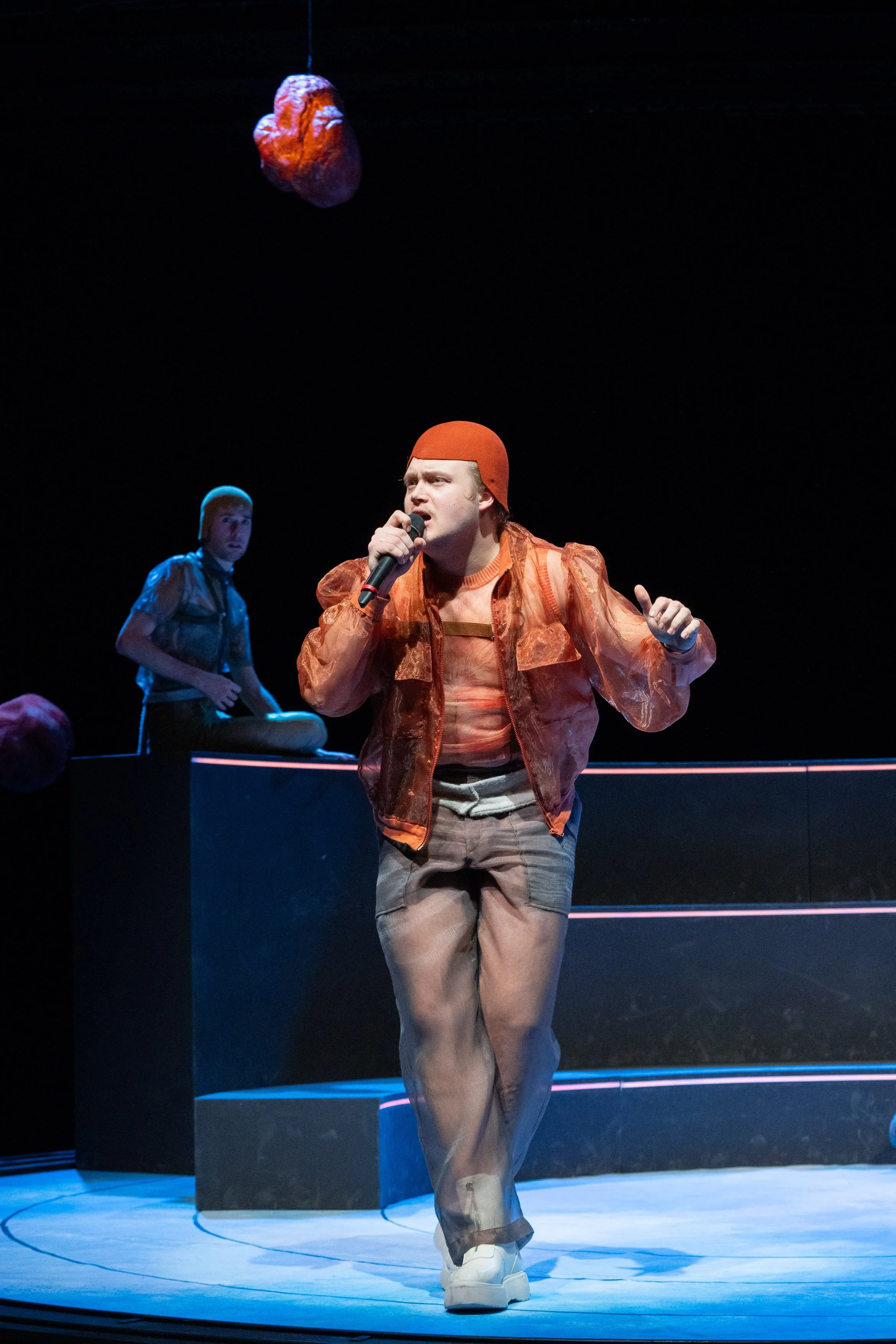 Schauspieler auf einer Bühne, der in der Hand ein Mikrofon hält und in einer roten Jacke, einem roten Hut und hellen Schuhen gekleidet ist, während im Hintergrund eine dunkle Szene mit Sitzen und another Schauspieler zu sehen ist.