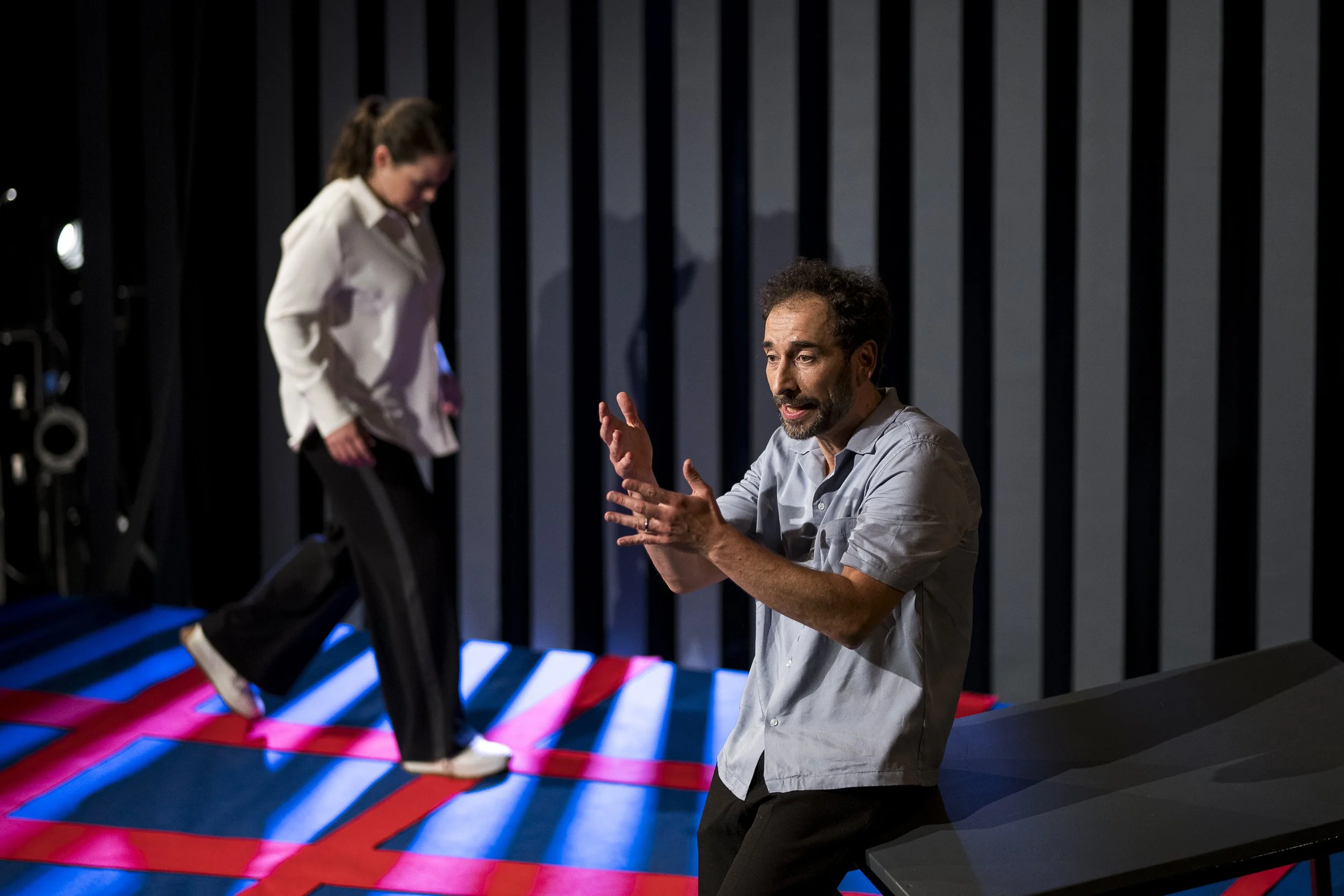 Ein Mann erklärt auf einer Bühne, während eine Frau auf einem Trampolin im Hintergrund springt.