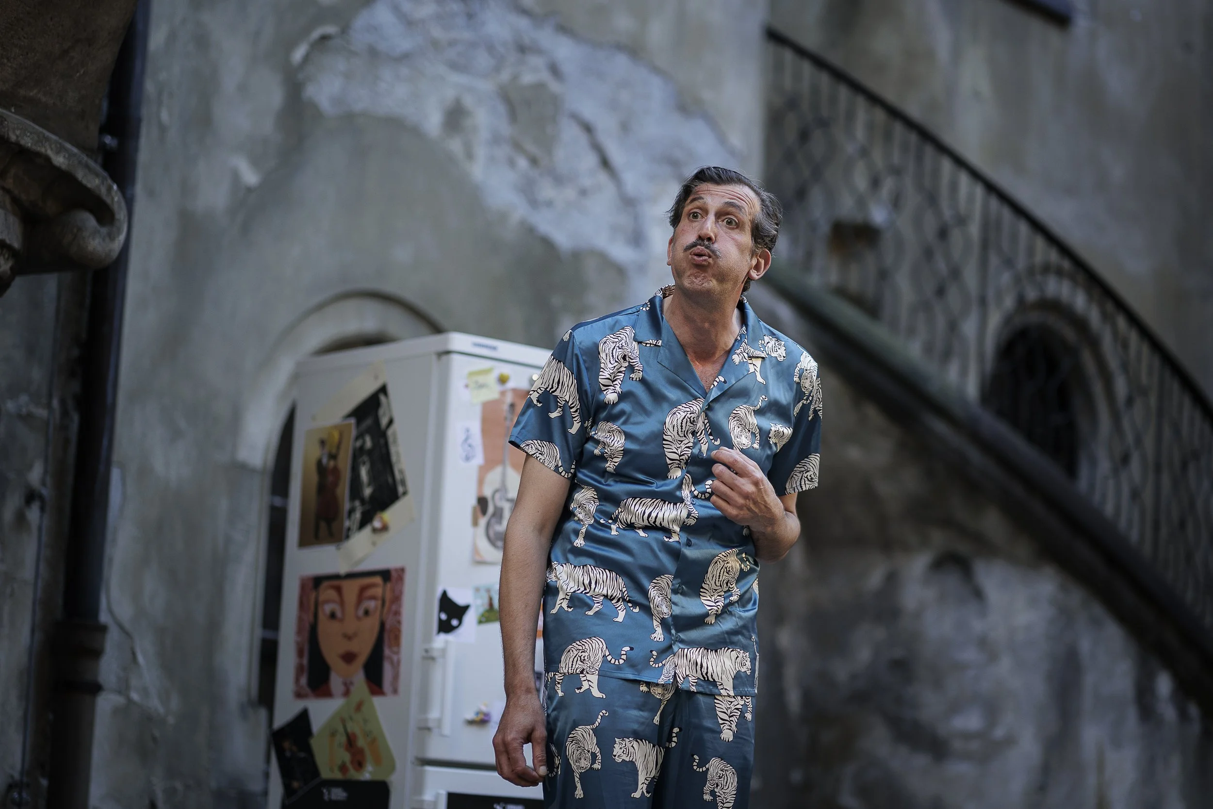 Ein Mann trägt ein blaues Hemd und Hosen mit Tiger-Motiv, steht in einem Raum mit einer alten, beschädigten Wand und einem weißen Kühlschrank mit Bildern und Post-its, während er eine nachdenkliche Pose einnimmt.