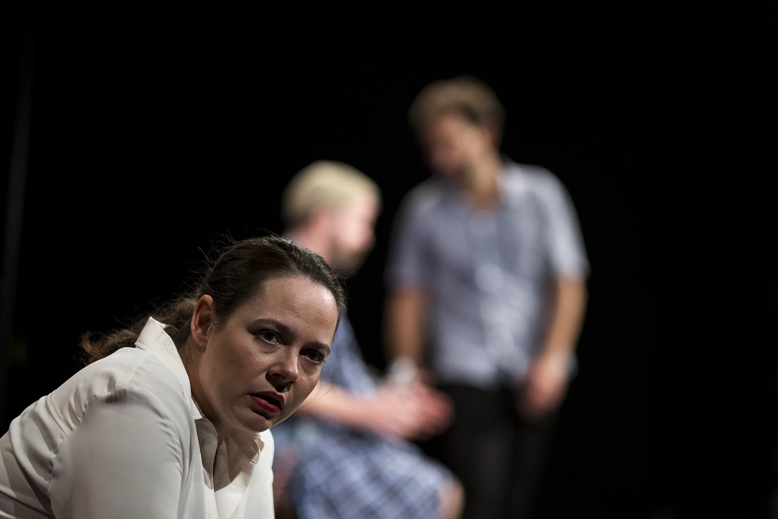 Drei Frauen auf einer Bühne, eine Frau im Vordergrund schaut ernsthaft, die zwei im Hintergrund sind unscharf, eine sitzt, die andere steht.