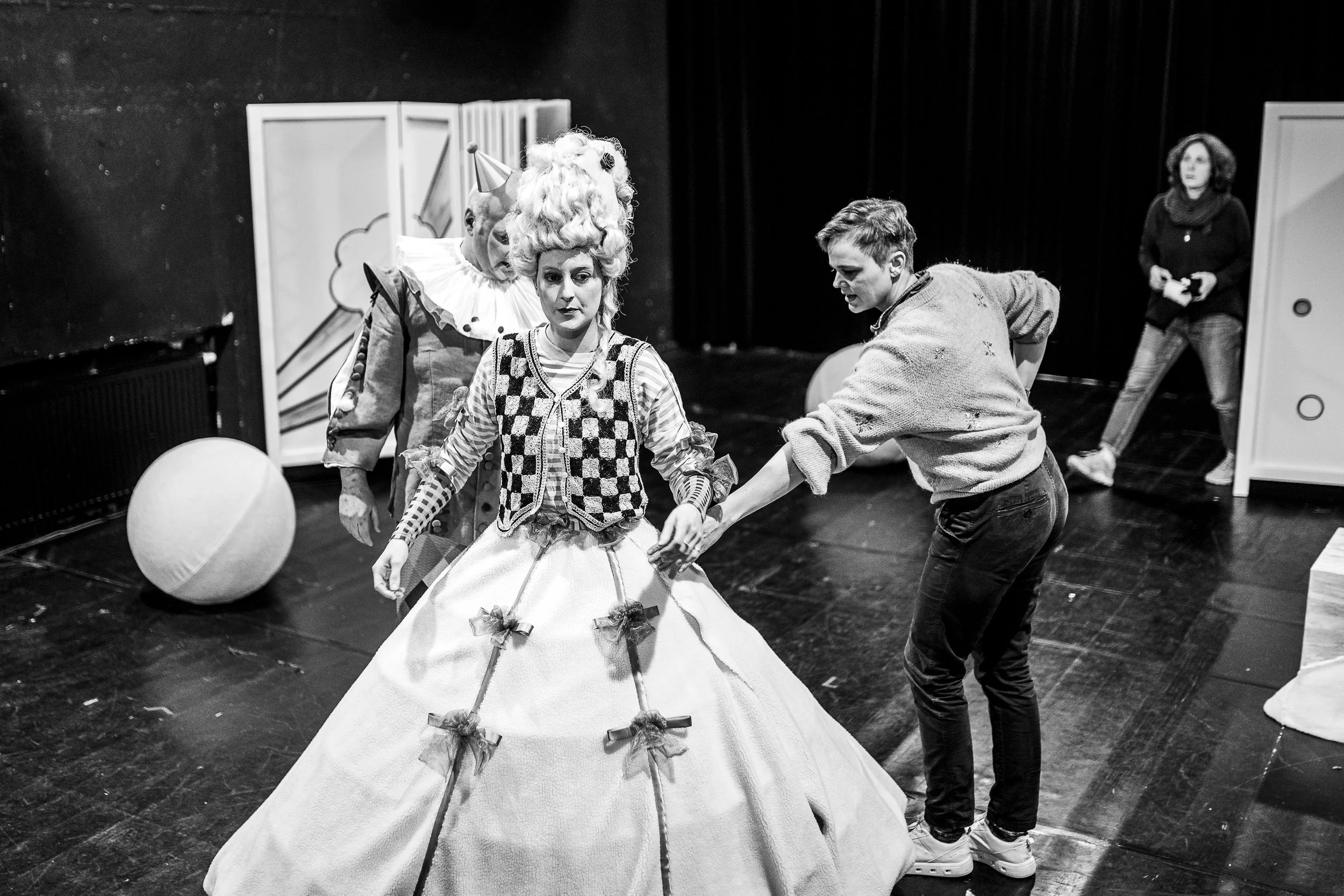 Drei Menschen auf einer Theaterbühne, einer trägt ein Kostüm und eine Perücke, was auf eine Theaterprobe oder Aufführung hinweist, im Hintergrund eine Frau beobachtet.