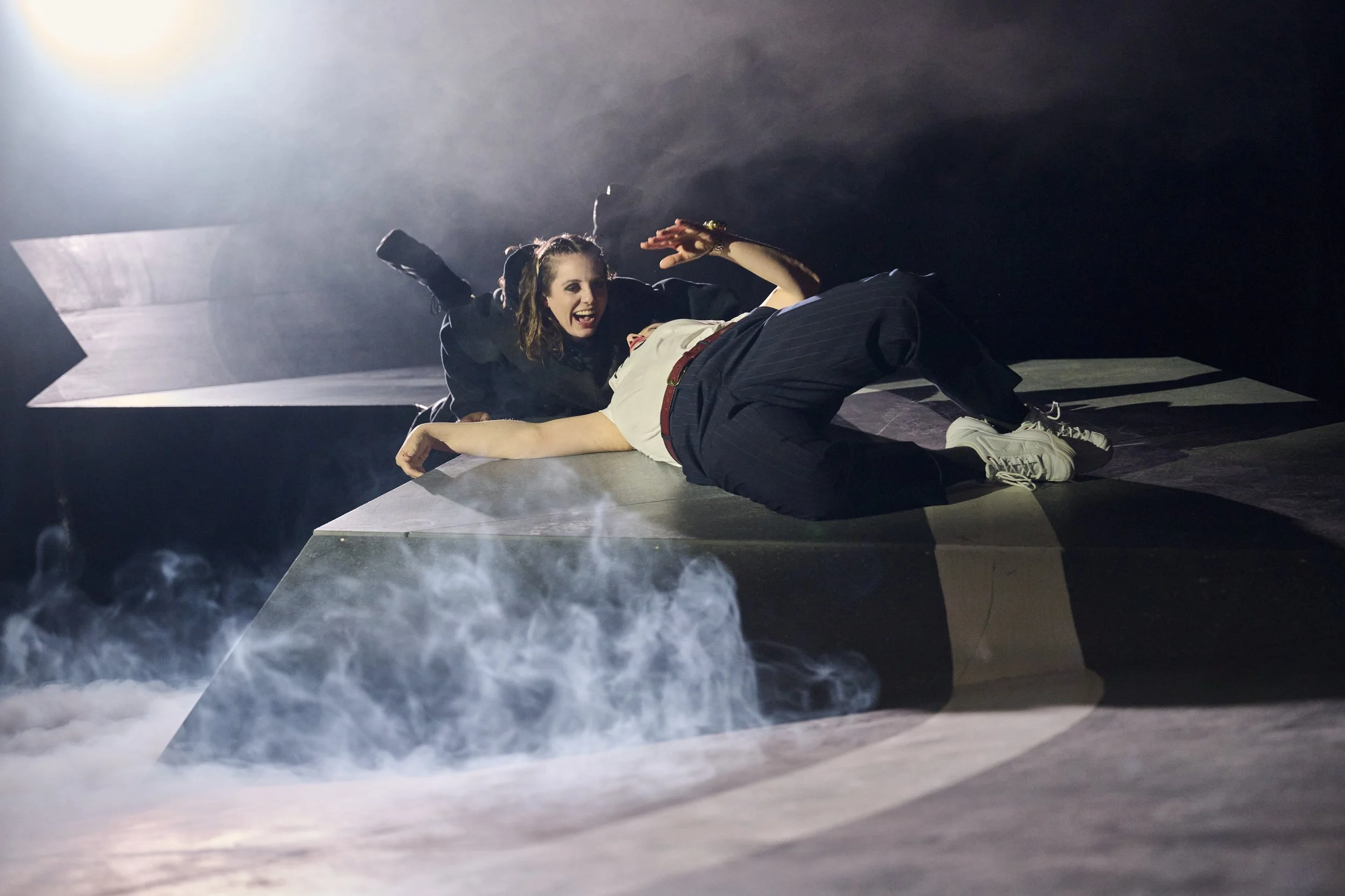 Zwei Frauen befinden sich auf einer surrealen schwarzen Bühne mit Nebel. Eine Frau liegt auf dem Boden und eine andere ist auf ihr und wirkt dramatisch, als ob sie eine Szene aus einem Theaterstück kommt. Peer Gynt (she/her) von Maria Milisavjevic.