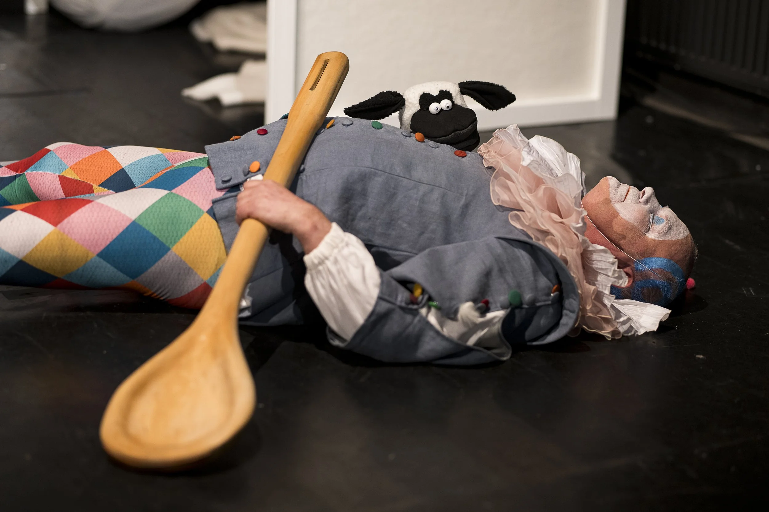 Ein Kosaken-Kriegsherr in einer Theaterproduktion liegt auf dem Boden, trägt eine Krawatte und eine graue Jacke, mit einer bunten, argyle-gemusterten Hose, und hält einen Holzlöffel in der Hand. Im Hintergrund ist eine Puppenfigur eines Hundes mit gr