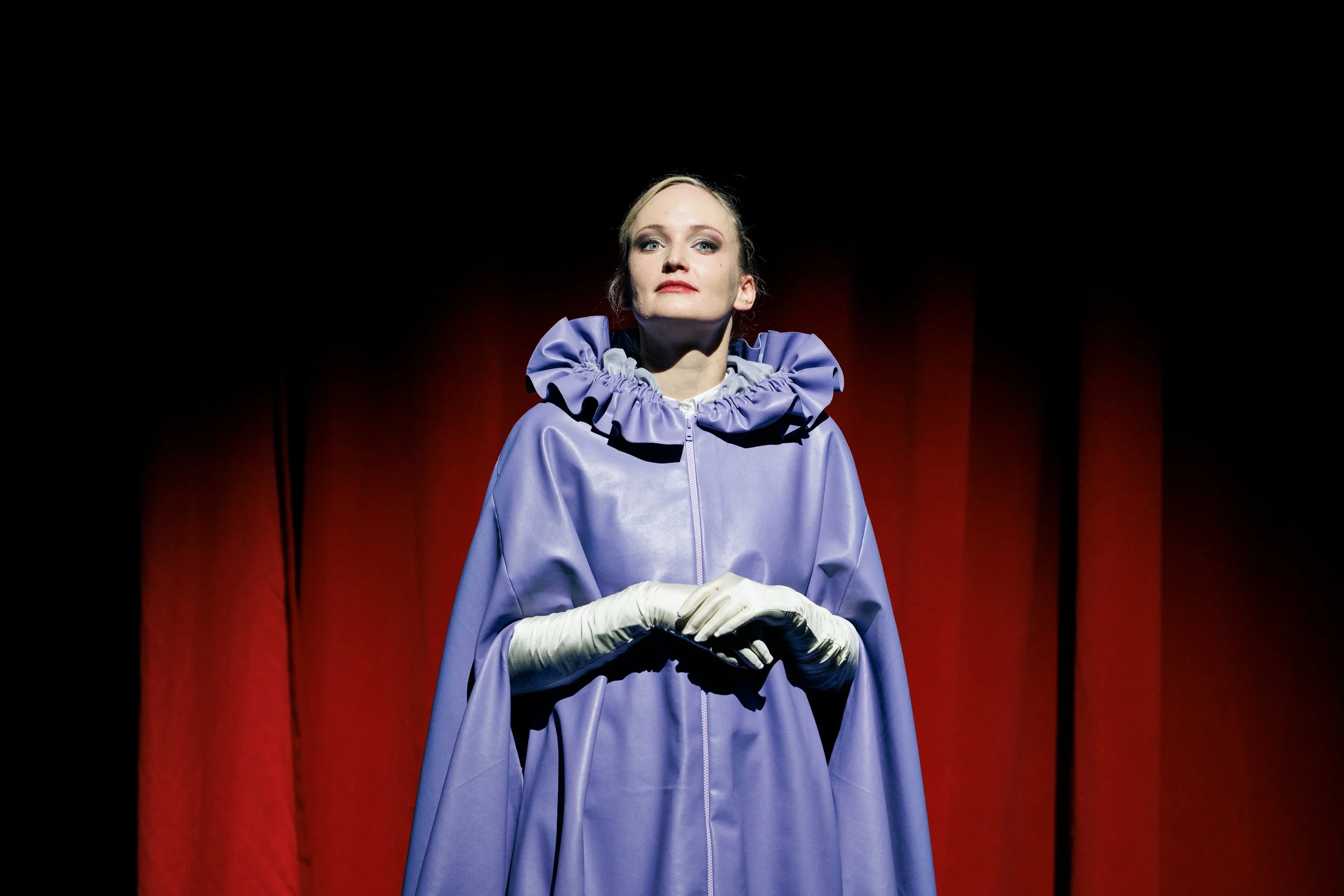 Eine Frau steht vor einem dunklen Vorhang, trägt einen lilafarbenen Umhang mit Rüschen am Hals und weiße Handschuhe.