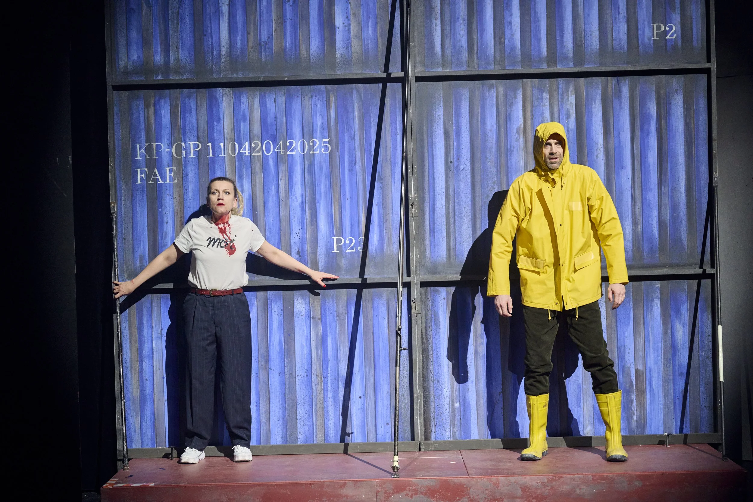 Zwei Schauspieler auf einer Bühne, eine Frau in lässiger Kleidung mit weißem T-Shirt und Theaterblut, ein Mann in einer gelben Regenjacke und Stiefeln, vor einer blauen Metallwand. Es könnte ein Container sein. 