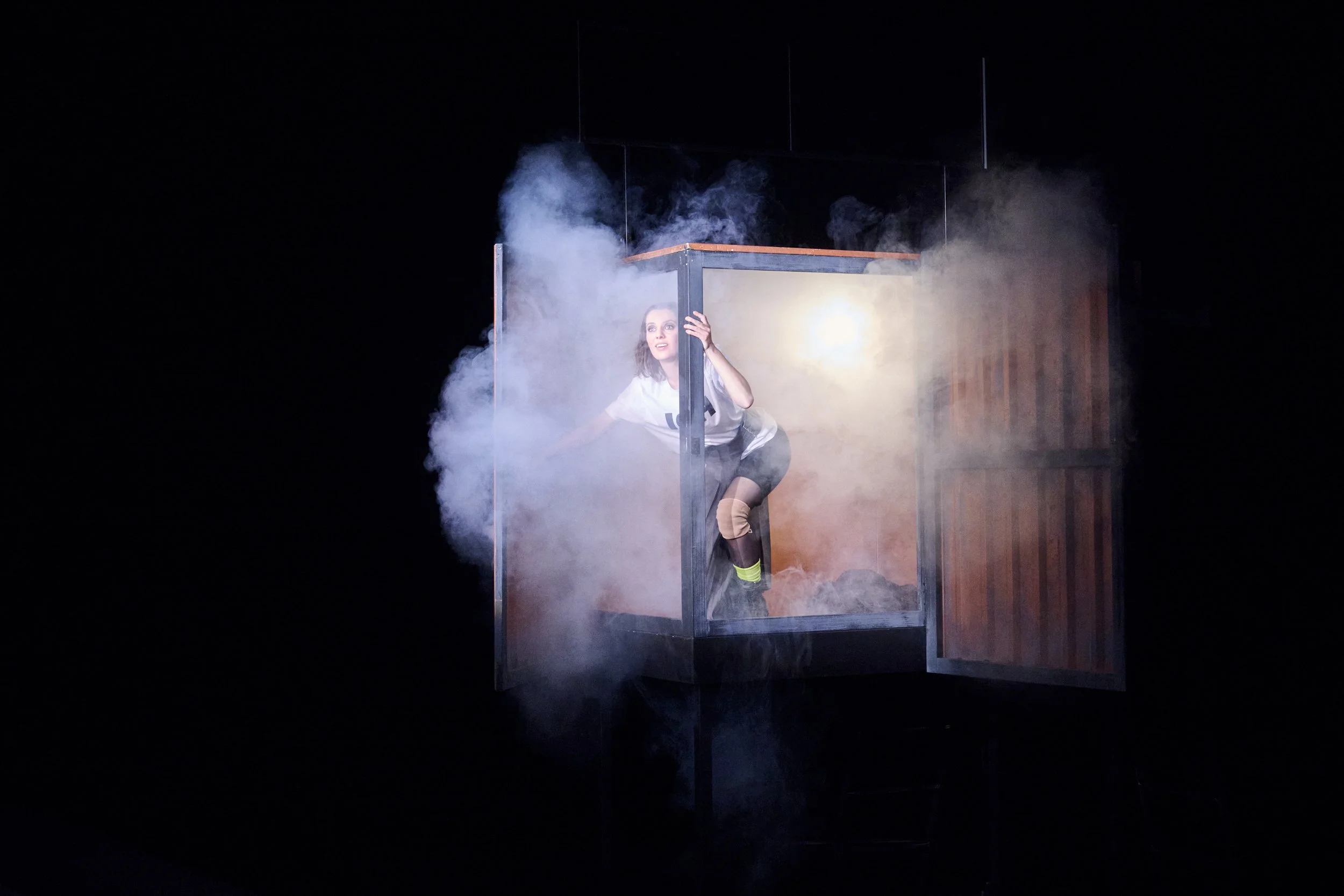 Eine Frau in einem schwarzen Body und weißen T-Shirt, die in einer Rauch- und Nebellandschaft auf einer Bühne in einem Holzrahmen gefangen ist.