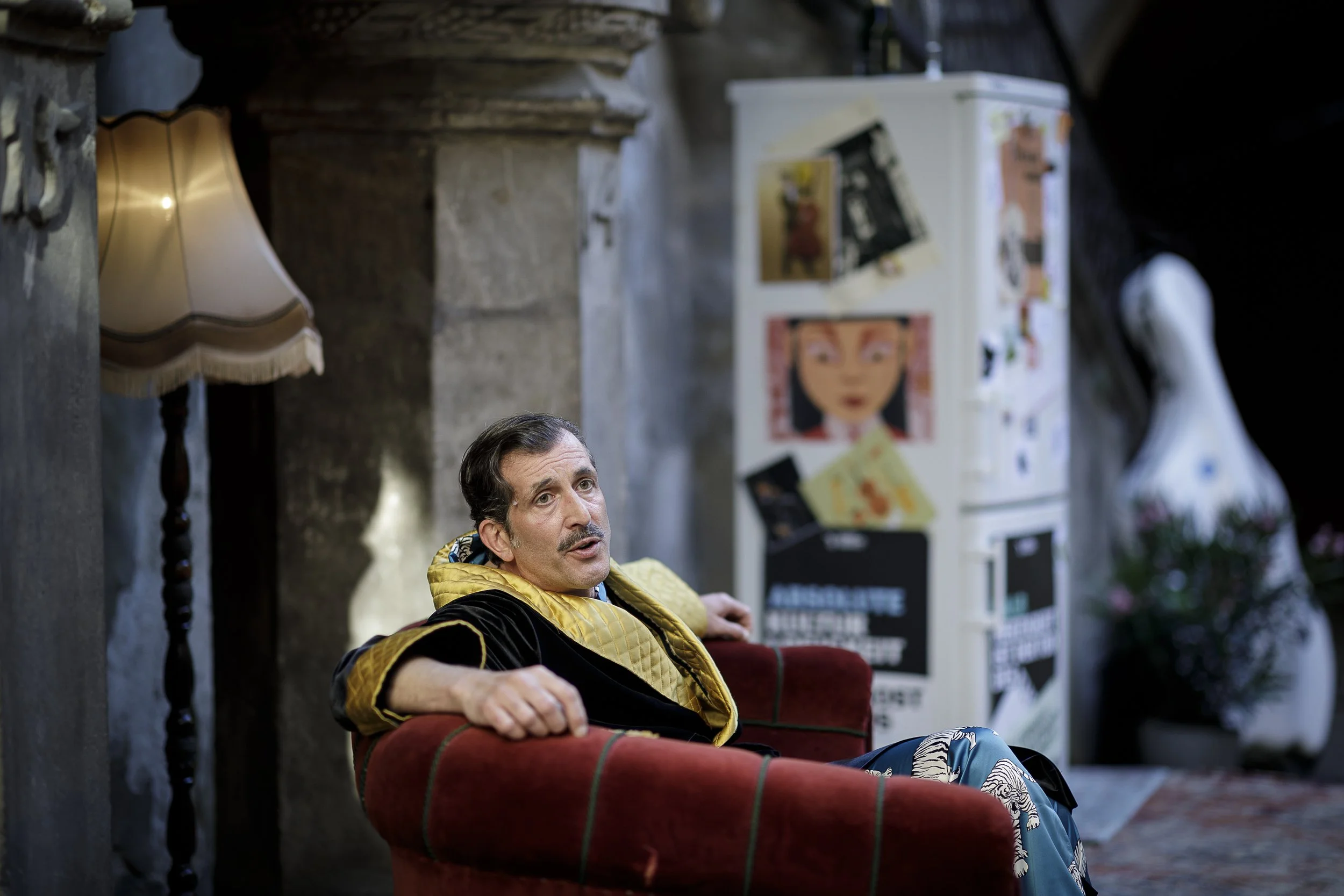 Ein Mann sitzt entspannt in einem roten Sessel, trägt einen schwarzen Mantel mit goldener Innenseite und blaue Hosen mit weißen Mustern, in einem gemütlichen Raum mit altem Steinmauer und einer Stehlampe. Im Hintergrund stehen Poster an einer weißen 