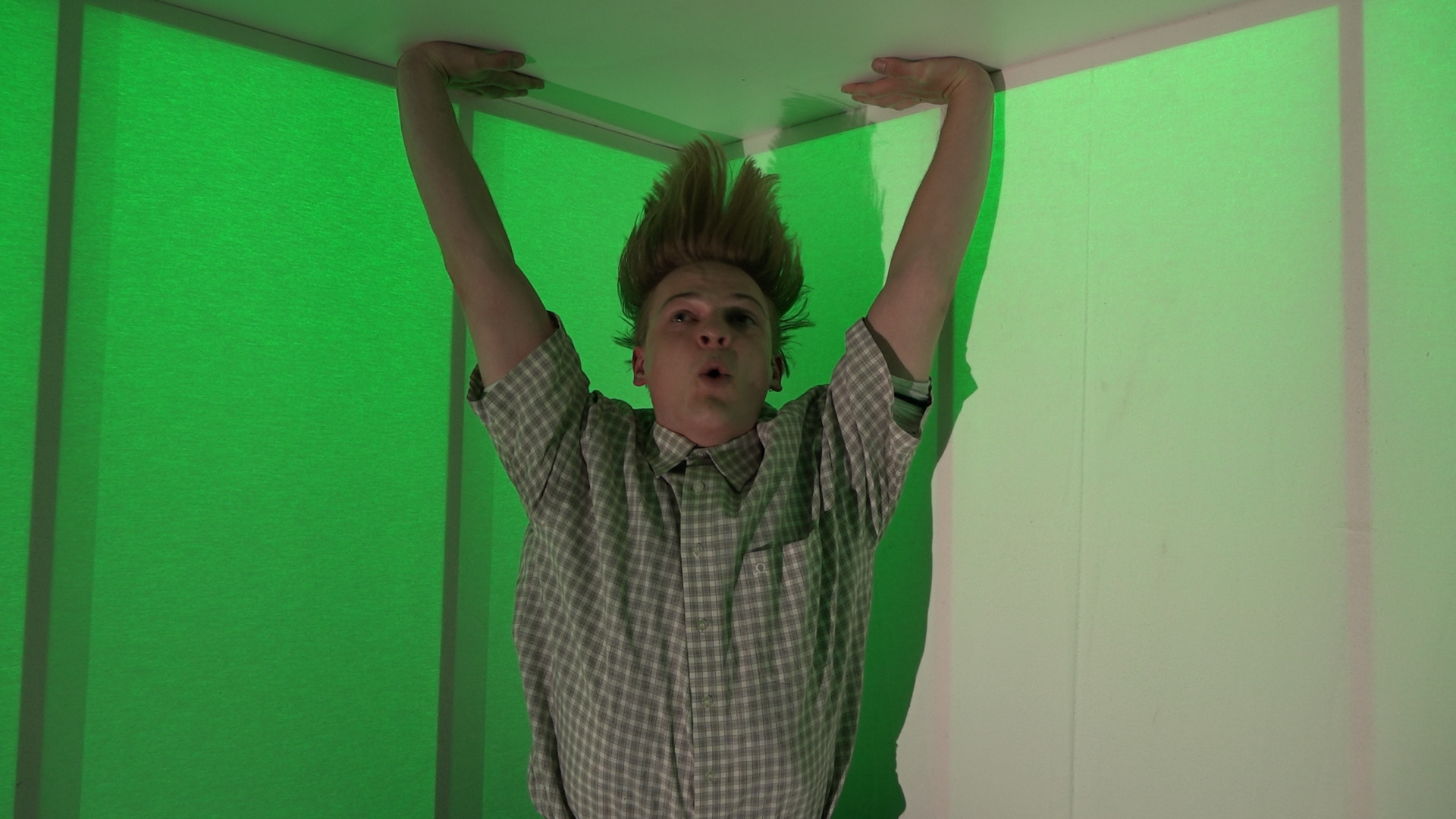 Junge Person mit aufgewirbelten Haaren, die an der Decke eines Raumes hängt, mit grüner Beleuchtung im Hintergrund.