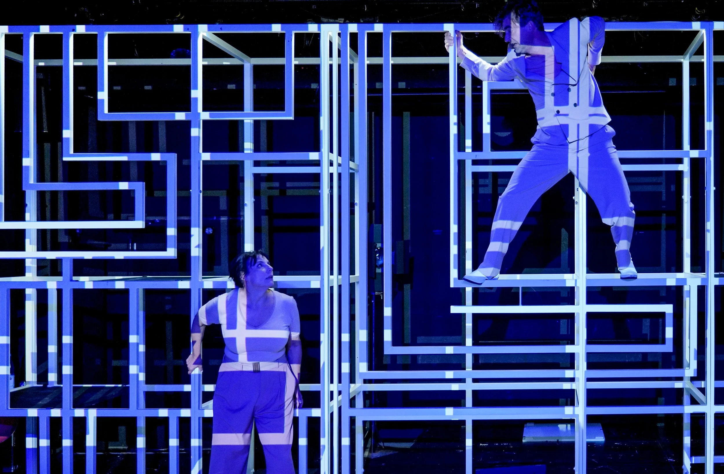 Zwei Personen in einem Theaterstück, die auf einer Bühne mit einer geometrischen Kastenstruktur stehen, die mit blauen Lichtmustern beleuchtet ist. Eine Person steht unten und schaut nach oben, während die andere Person oben im Kasten steht und in ei