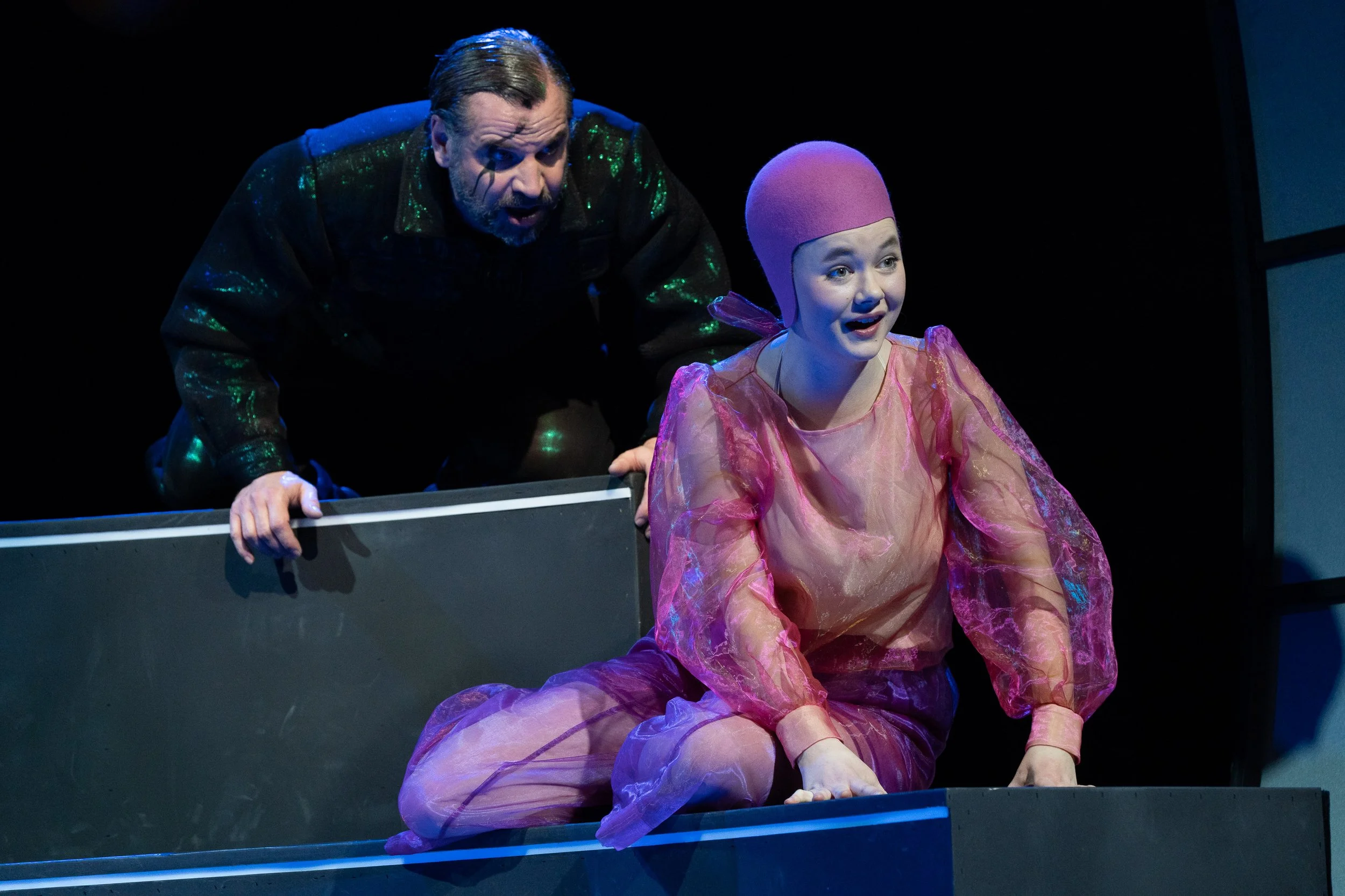 Zwei Schauspieler in einem Theaterstück, einer steht im Hintergrund, der andere sitzt vorne, beide in farbenfrohen Kostümen.