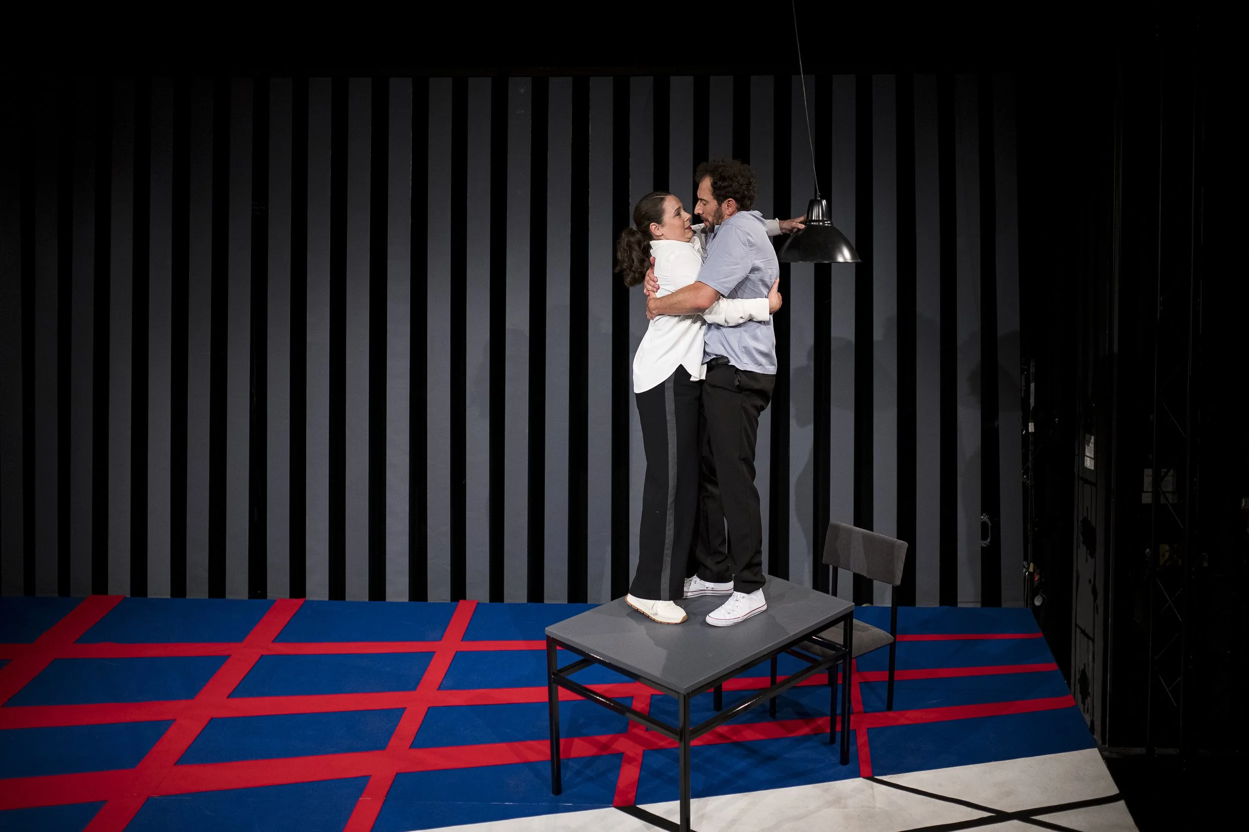 Zwei Schauspieler stehen auf einem Tisch, umarmt und schauen sich tief in die Augen, in einer Theaterbühne. Hinter ihnen eine schwarze Wand mit vertikalen Linien, auf dem Boden ein rotes und blauer Musterteppich.