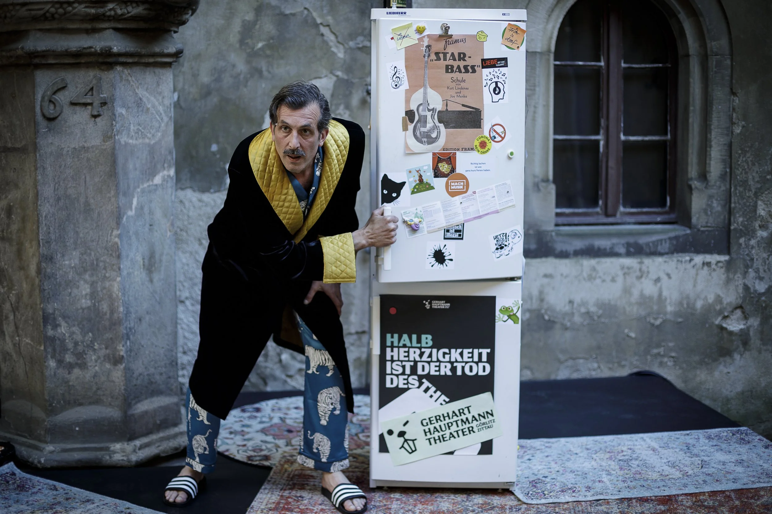 Ein Mann in einem schwarzen Bademantel mit gelben Quilted-Details und blauen Haremshosen mit Eulenmuster steht neben einem Whiteboard, das mit verschiedenen Postern, Notizen und Stickern bedeckt ist. Er trägt schwarze und weiße Adidas-Schlappen und b