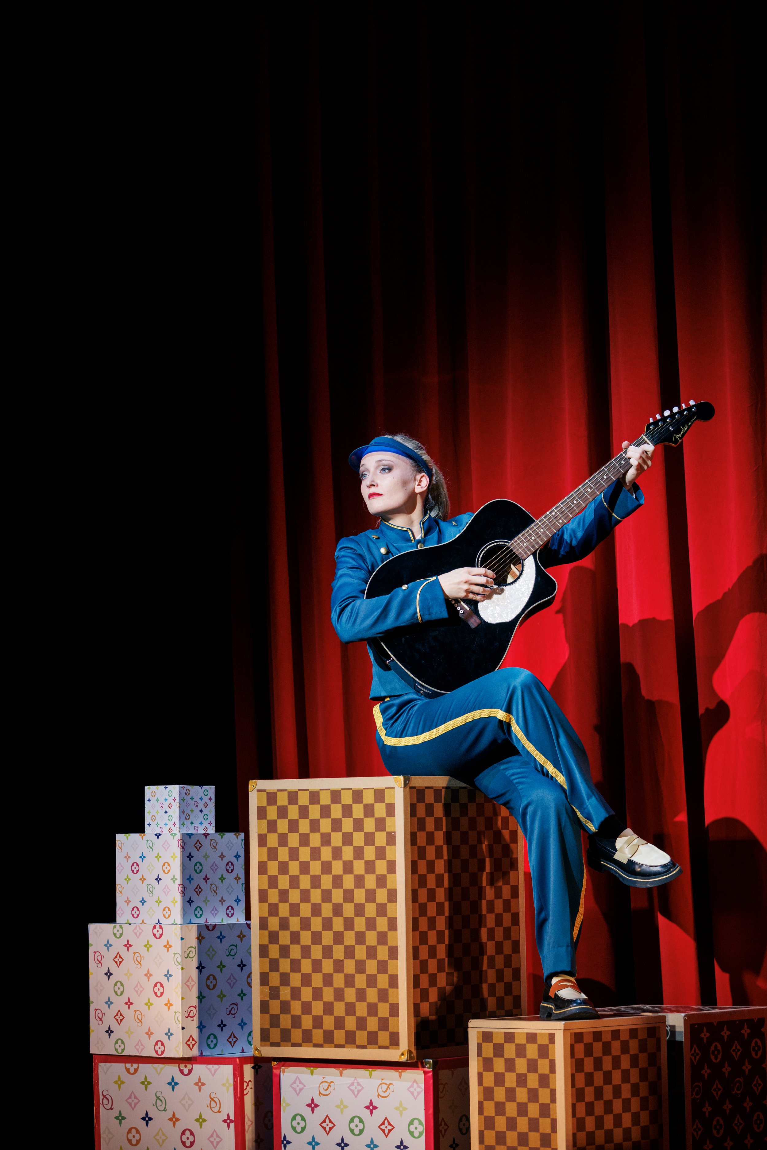 Eine Frau in blauer Kostüm auf einer Bühne, sitzend auf braunen Kisten, spielt Gitarre vor roten Vorhängen.