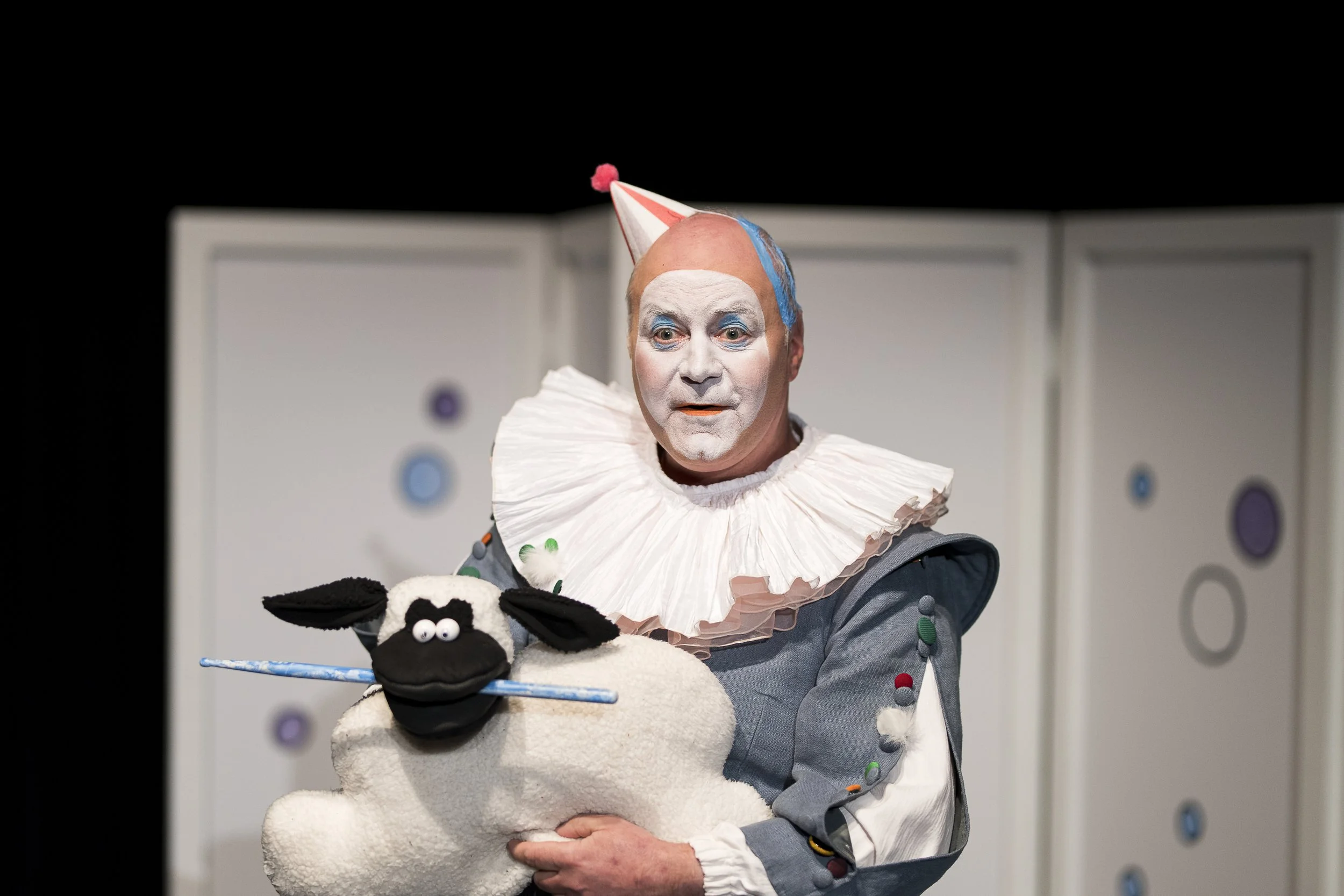 Ein Clown mit weißem Gesicht, buntem Hut und Rüschenkragen hält eine Schaf-Puppe auf der Bühne. Er trägt ein graues Kostüm mit bunten Pompons und hat einen überraschten Ausdruck. Im Hintergrund sind weiße Kulissen mit bunten Kreisen.