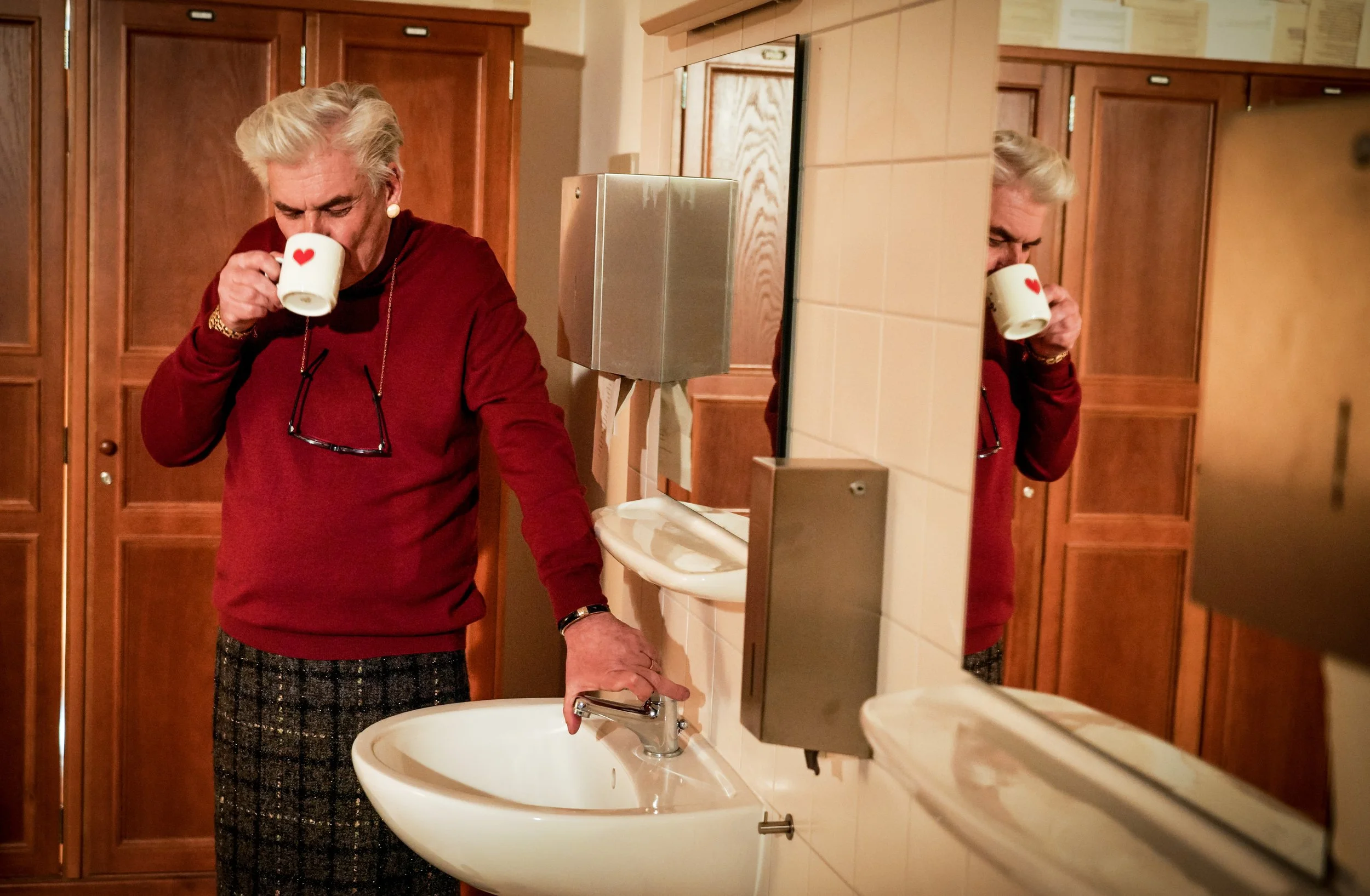 Ältere Frau mit Grauem Haar trinkt Kaffee aus einer weißen Tasse mit rotem Herz, steht vor einem Waschbecken in einer Toilette, trägt eine rote Jacke, eine dunkle Brille und hat eine Armbanduhr sowie eine Halskette. Sie steht vor einem Wandspiegel, d