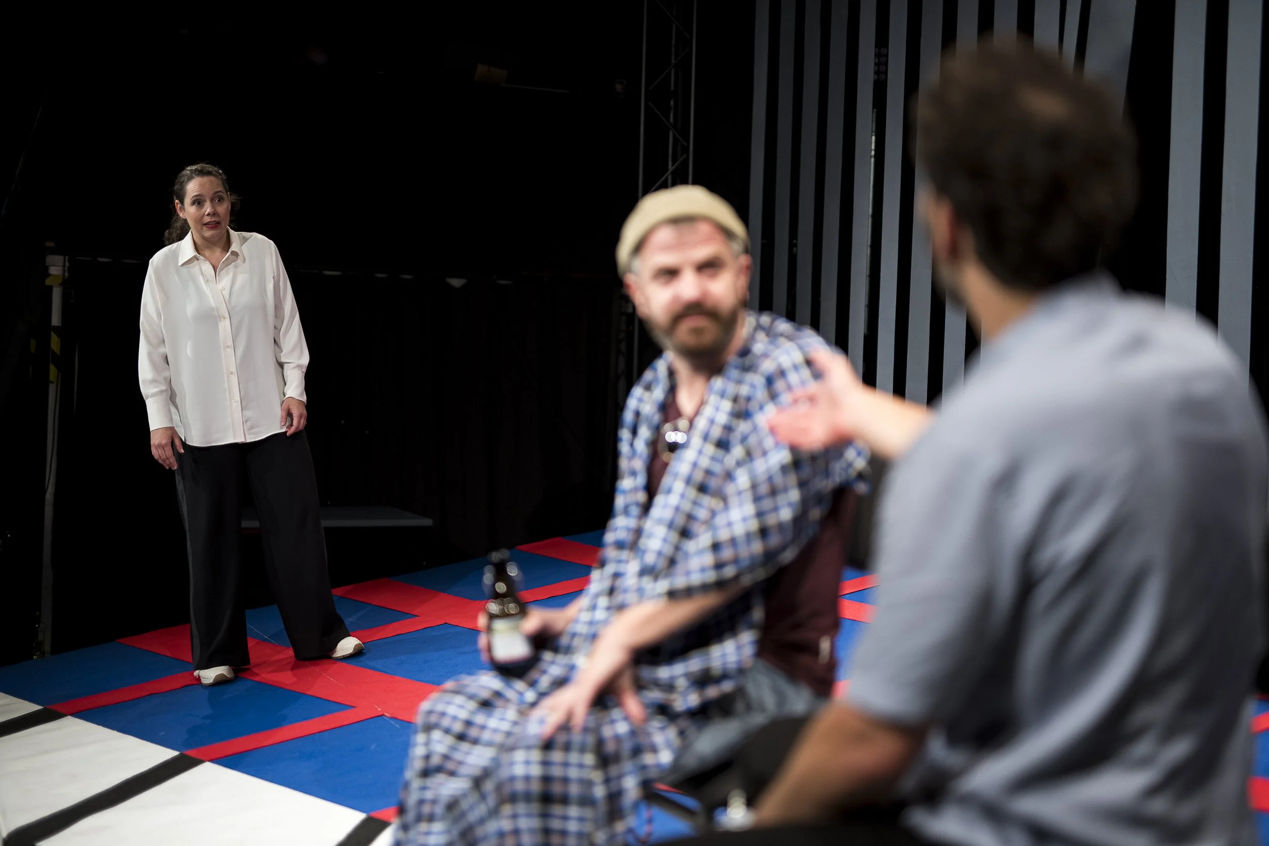 Dreipersonen-Theaterprobe mit Frau im weißen Hemd und zwei Männer in lockerer Kleidung auf einem bunten Bühnenboden