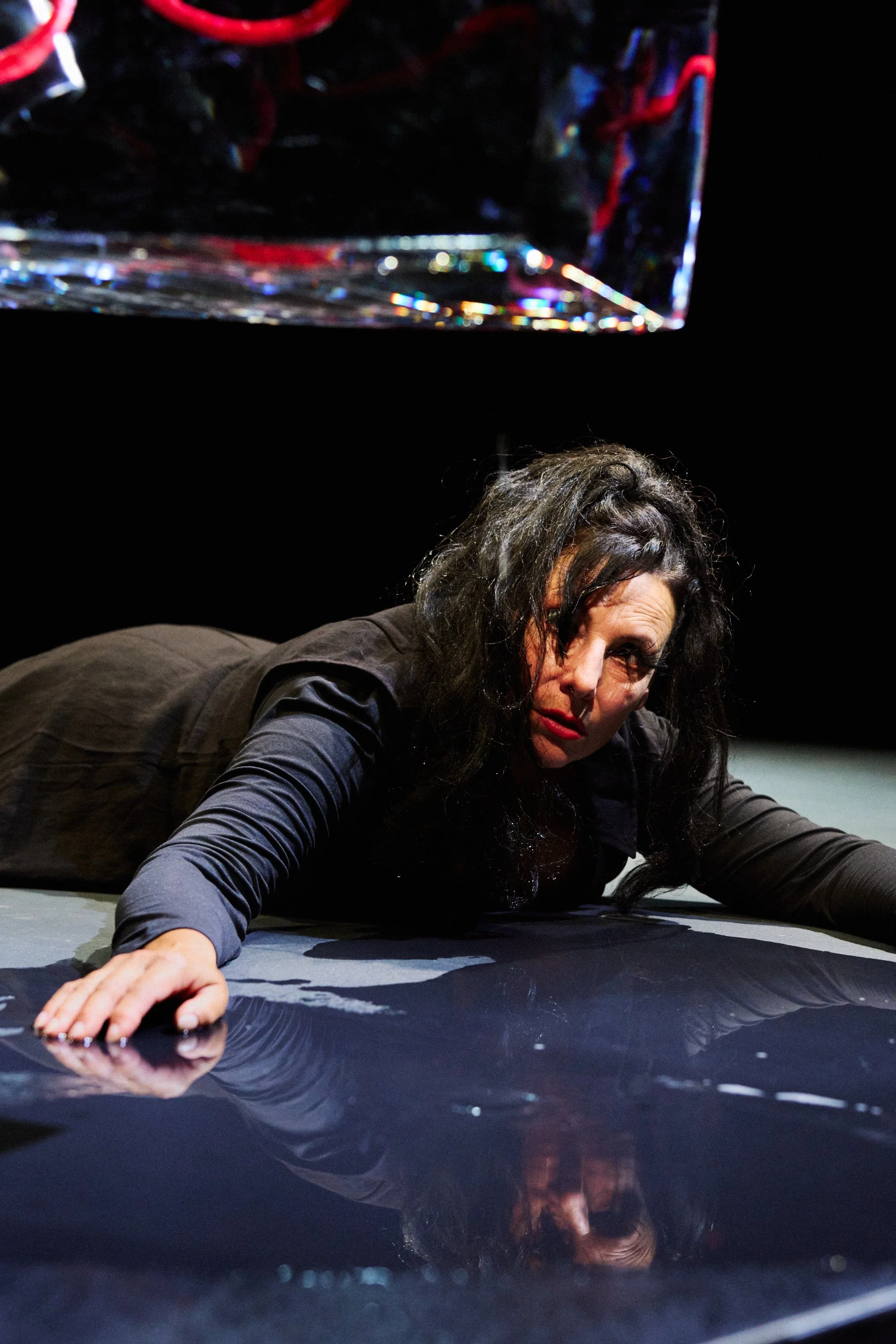 Frau liegt auf dem Boden, blickt erschöpft und konzentriert. Sie hat dunkle, zerzauste Haare und trägt dunkle Kleidung. Der Hintergrund ist schwarz, und es gibt eine reflektierende Oberfläche, auf der sie schaut.