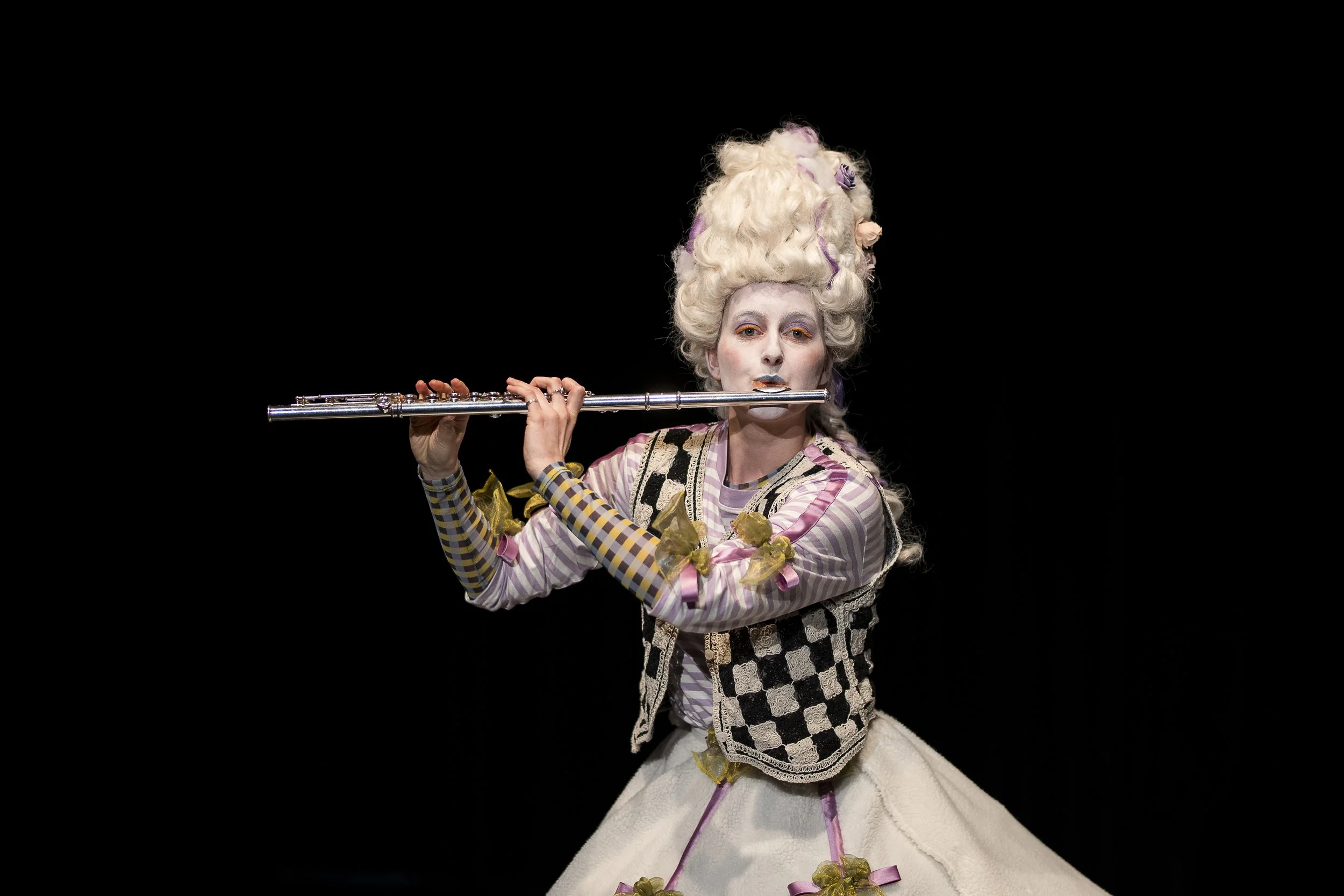 Theaterfigur mit weißem, aufwendig frisiertem Haar, trägt bunte Kleidung, spielt Flöte, vor schwarzem Hintergrund.