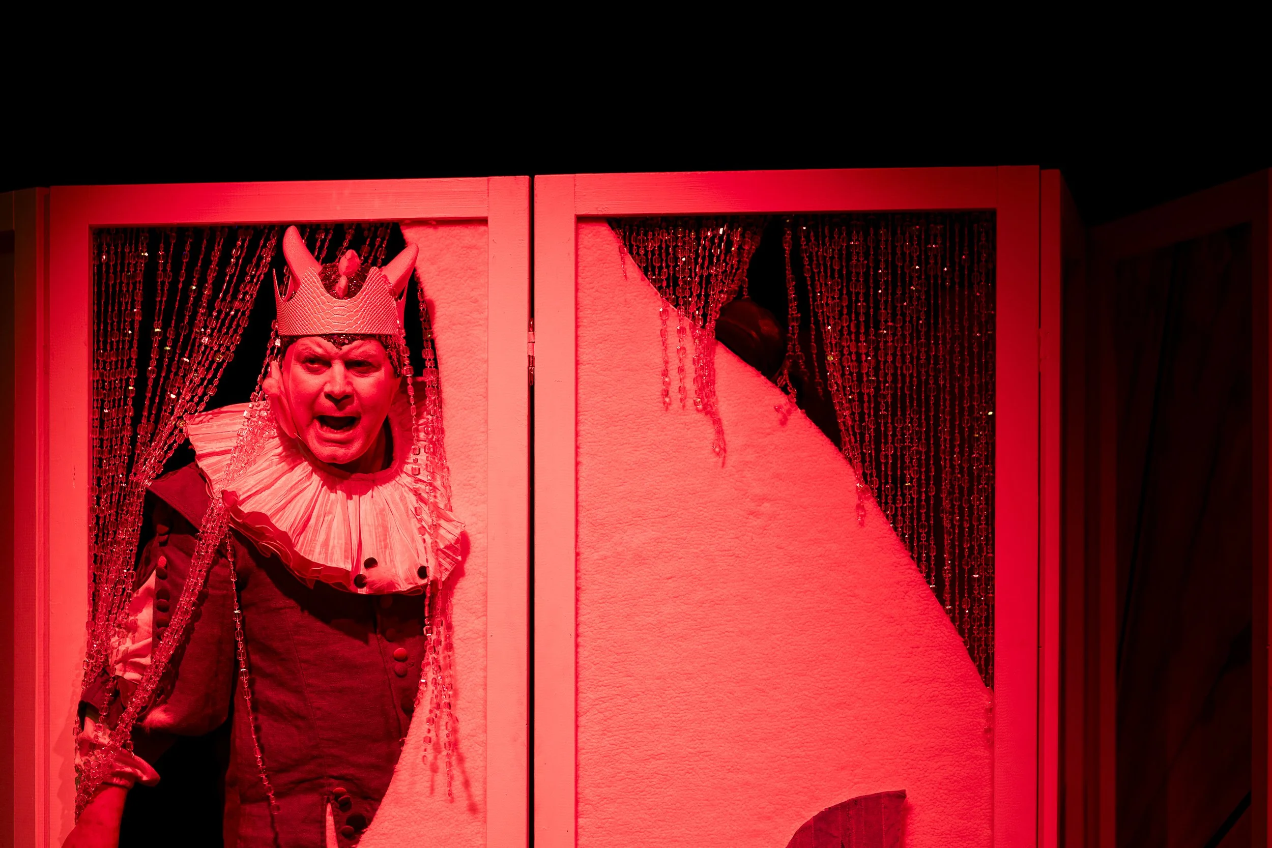 Ein Mann als Kasper, der aus einer im roten Licht getauchten Bühne schaut, trägt eine Krone und ein Kostüm mit großer Renaissance-Kragen, hinter roten Vorhängen.