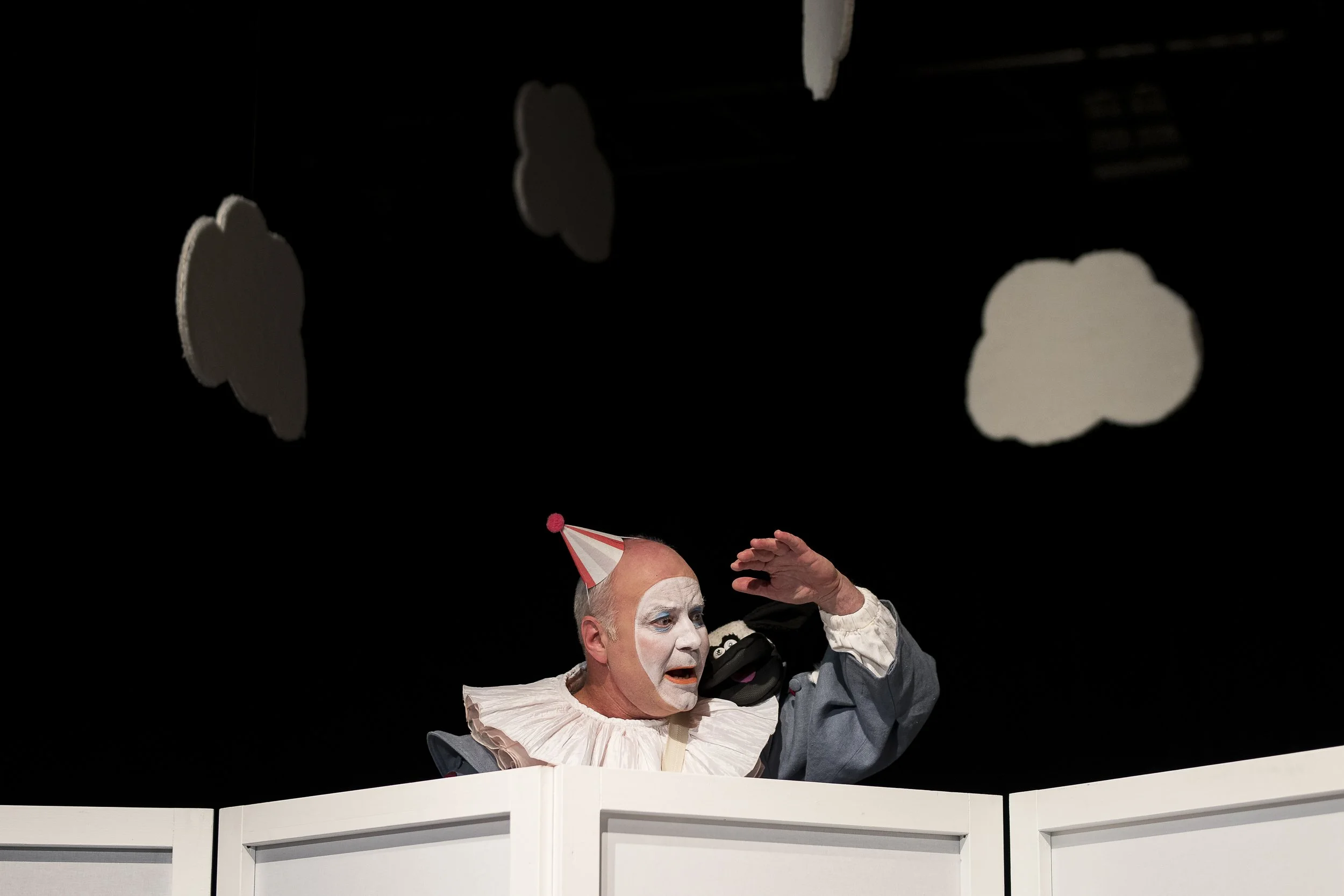 Clown mit weißem Gesicht, rotem Partyhut, grauem Kostüm und Rüschenkragen, schaut überrascht und weint, während er auf einer weißen Bühne steht, mit schwarzen Hängeschirmen und Wolken im Hintergrund.