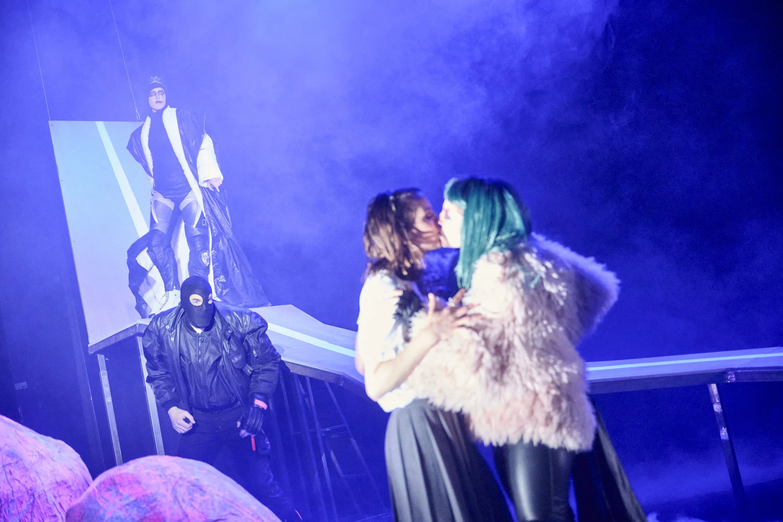 Zwei Frauen küssen sich auf einer Bühne, im Hintergrund sind zwei Personen in maskierten, dunklen Outfits, eine auf einer erhöhten Plattform, die andere am Boden, mit Nebel und blauem Licht.