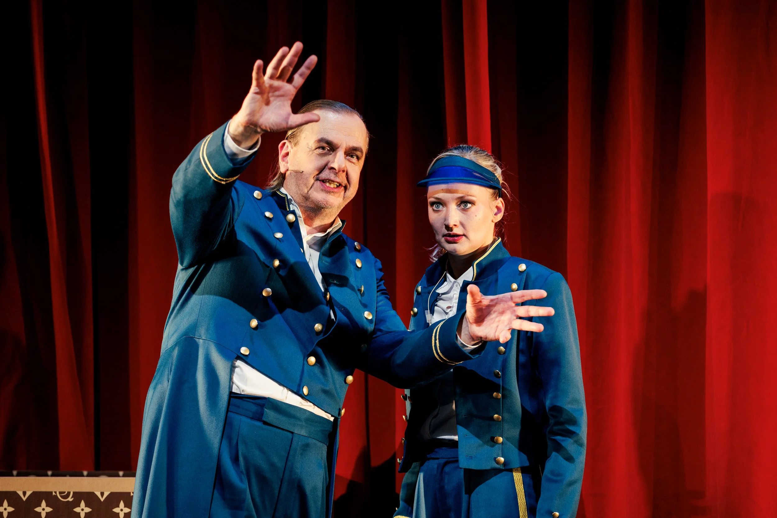 Zwei Schauspieler in blauen Uniformen auf einer Bühne vor roten Vorhängen, einer spricht mit Handbewegung, die andere schaut aufmerksam.