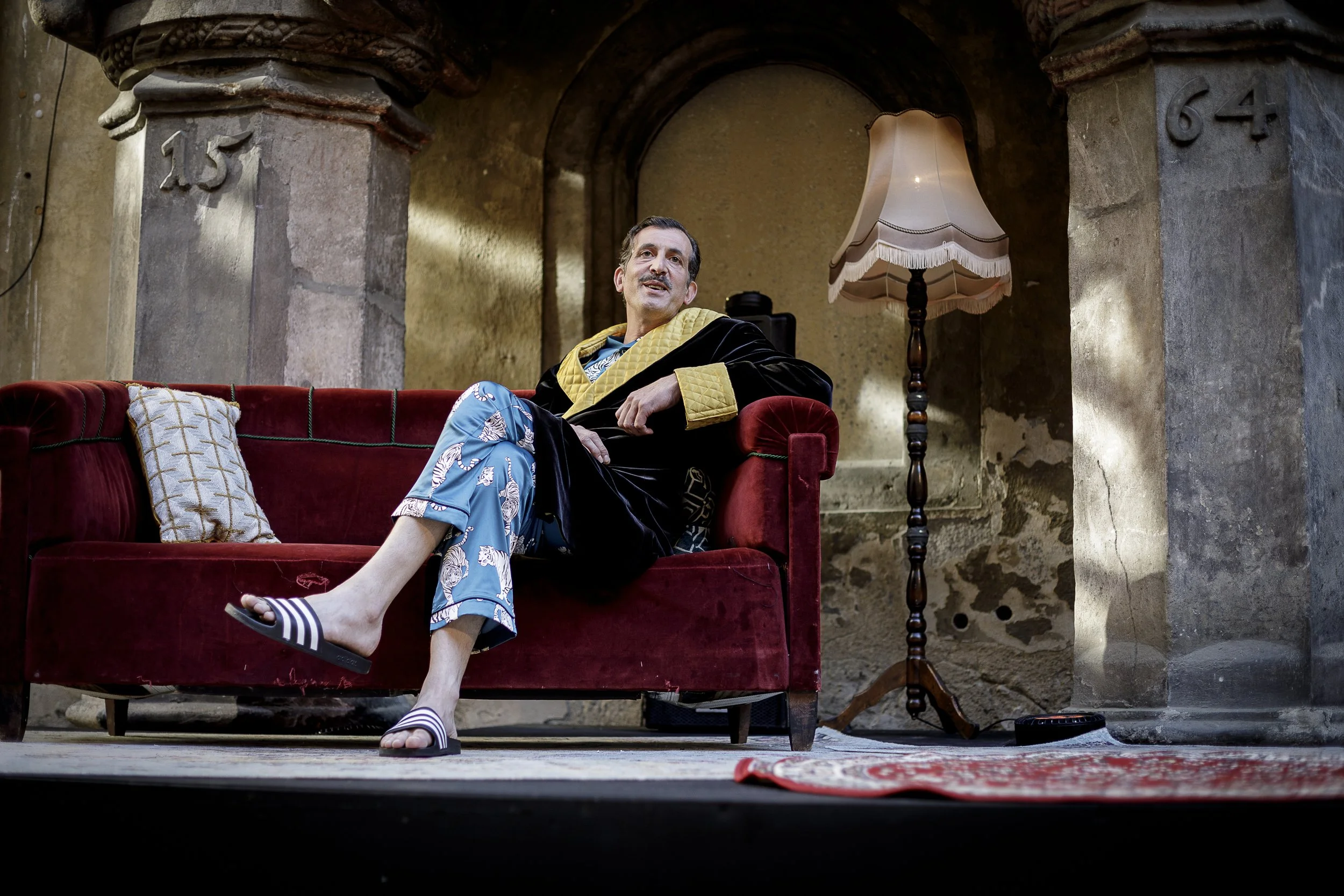 Mann sitzt entspannend auf einem roten Sofa in einem alten Raum mit abblätternder Wand, trägt eine schwarze Robe mit goldenen Akzenten und Pyjamahosen, trägt Slipper, neben einem alten Stehlampe, im Hintergrund die Nummern 25 und 64.