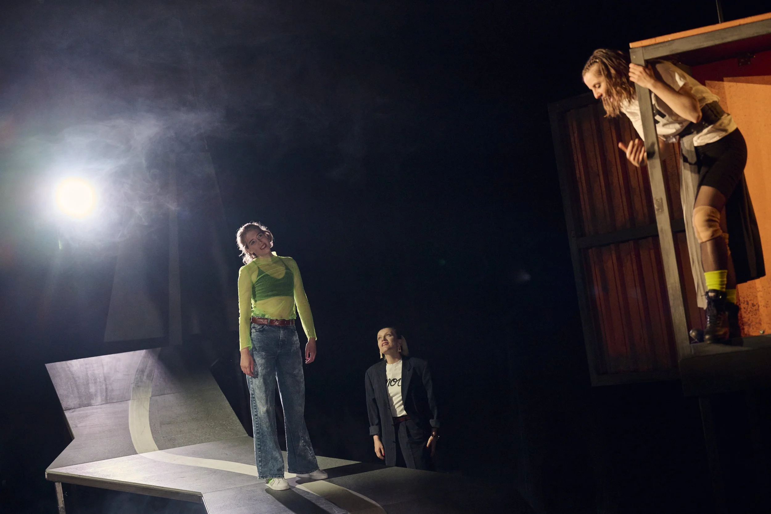 Drei junge Frauen auf einer Bühne, eine steht auf einer Art Strasse und eine andere steht hinter ihr aber auf dem Boden, während die dritte aus einem nebligen Holzcontainer lehnt, bei einer Theateraufführung bei Nacht. Gegenlicht und Nebel.