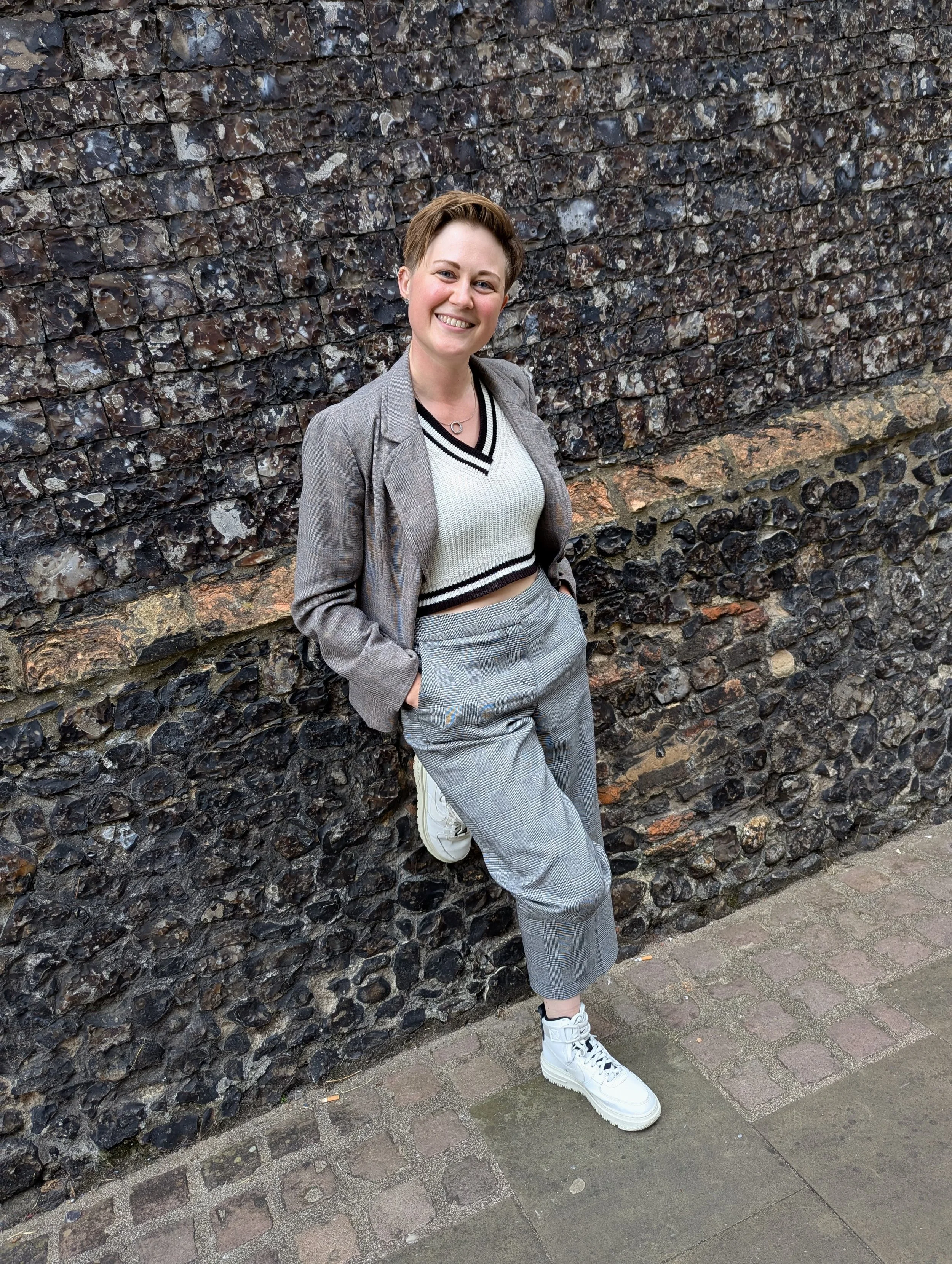 A young woman with short hair smiling, wearing a gray blazer, a white v-neck sweater, checkered pants, and white sneakers leaning against a stone wall with her hands in her pockets.