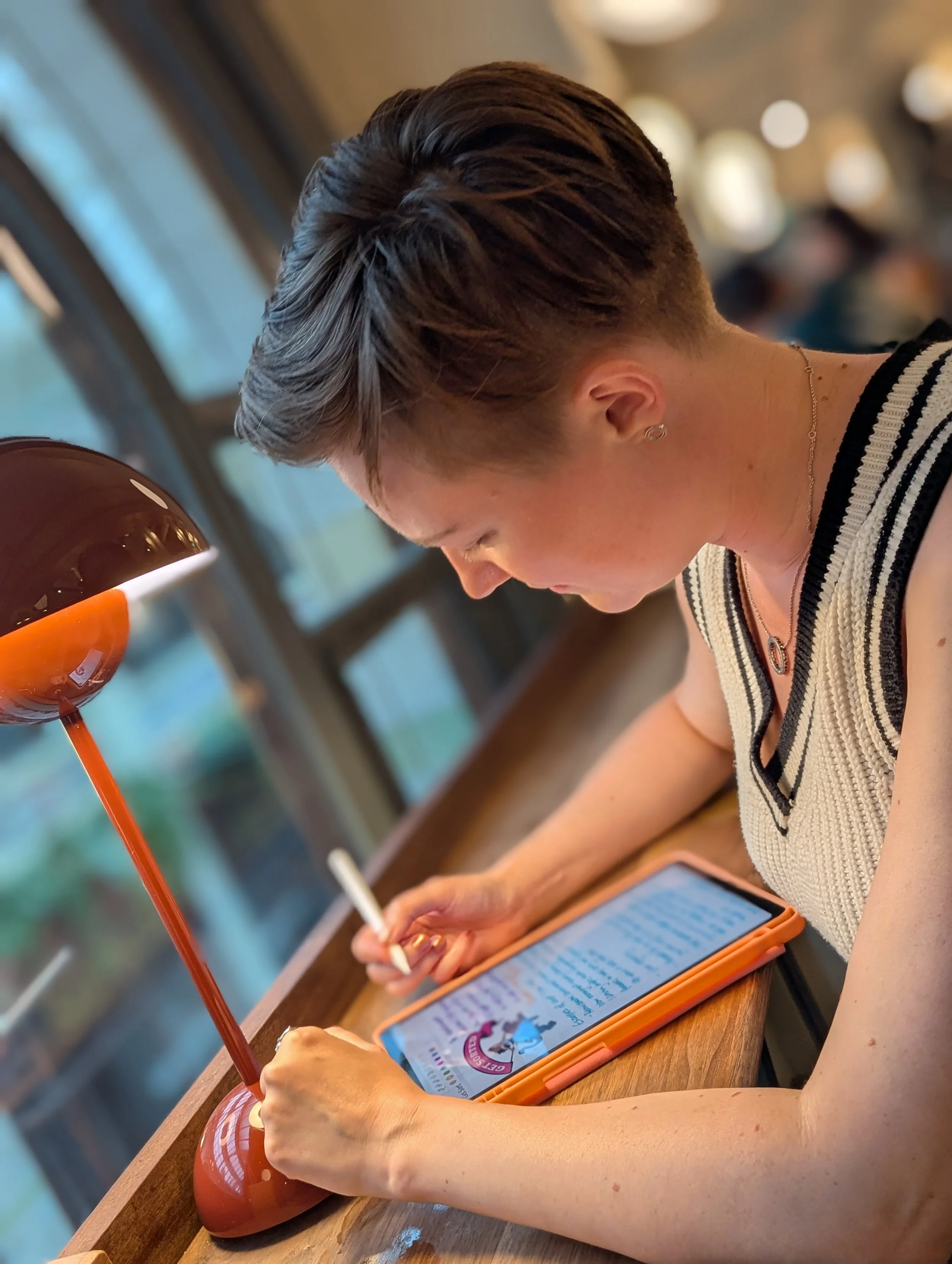 A woman with short, styled hair sitting at a wooden table, reading on a tablet with a stylus in a cozy indoor setting with warm lighting.