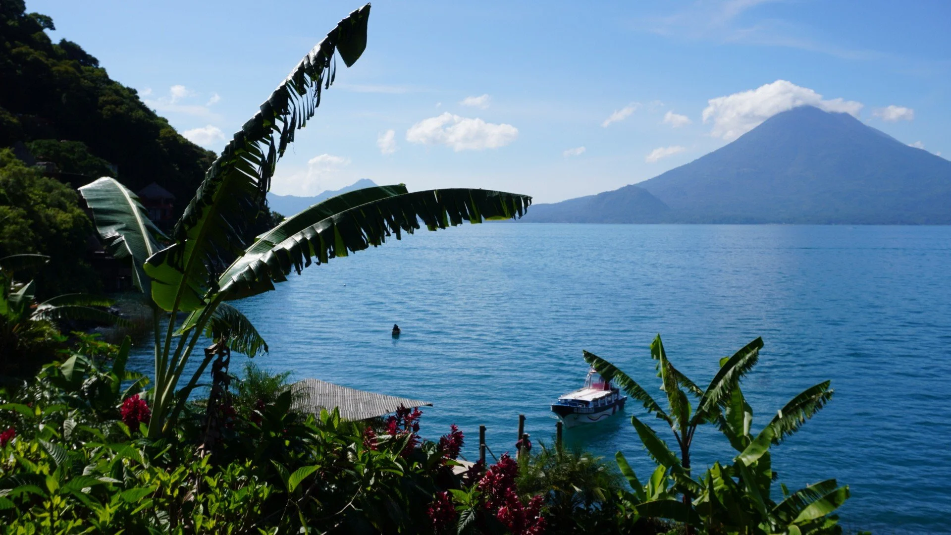 Tropical lake with lush greenery and banana plants in the foreground, a boat on the water, with volcanic mountains in the background under a partly cloudy sky.