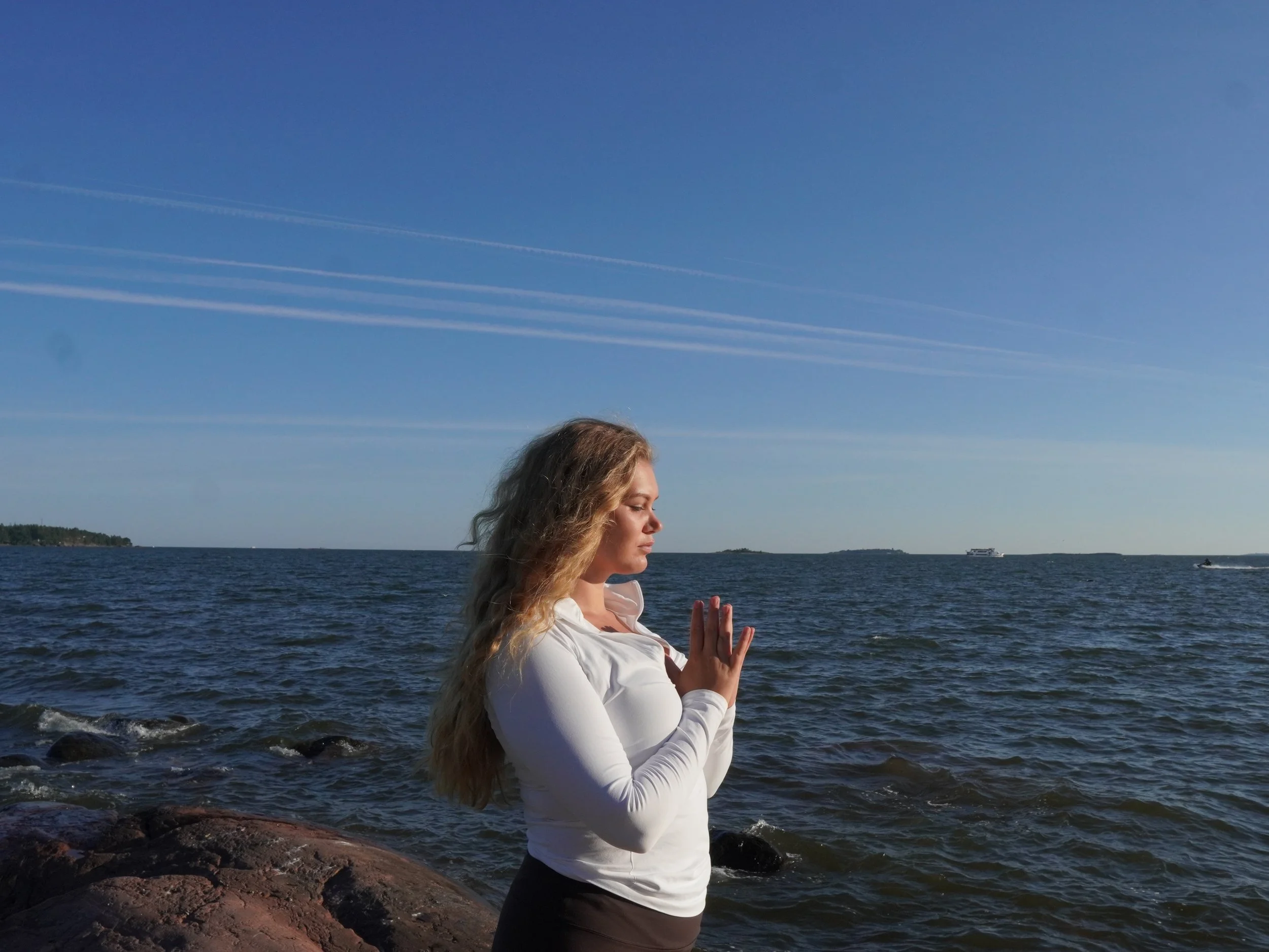 A woman practicing meditation or yoga by the water on a sunny day with a clear blue sky and contrails. She is in a meditative pose with hands in prayer position, eyes closed, standing on rocks at the shoreline.