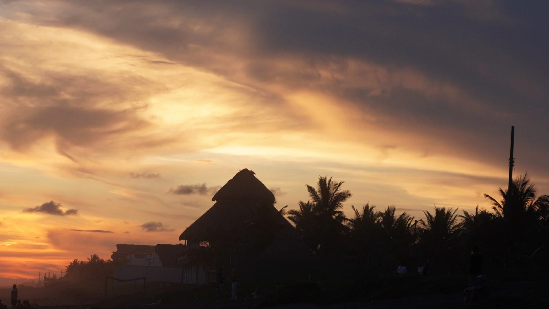 Sunset scene with a thatched-roof hut, palm trees, and a cloudy sky with warm golden hues.