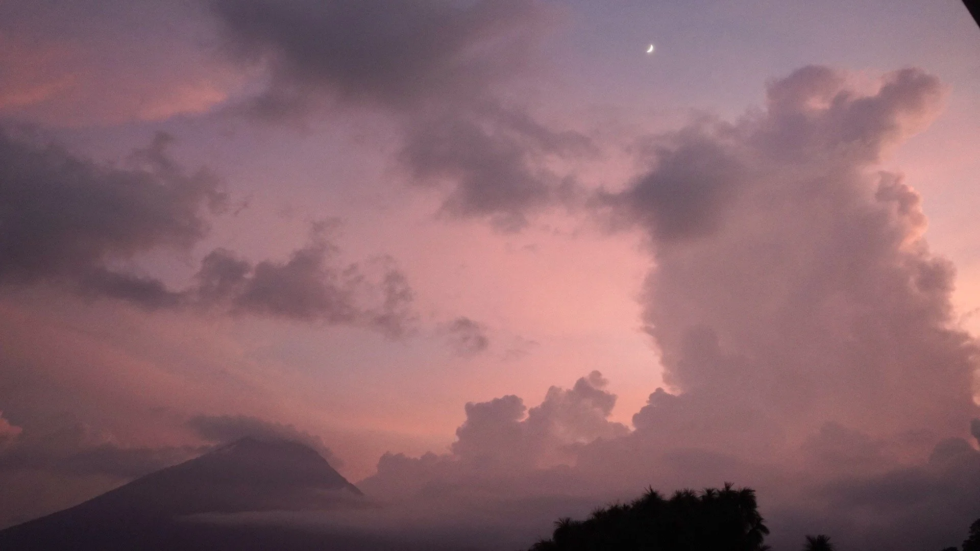 A scenic view of mountains at sunset at lake Atitlan, Guatemala, with dark clouds and a crescent moon in the purple and pink sky.
