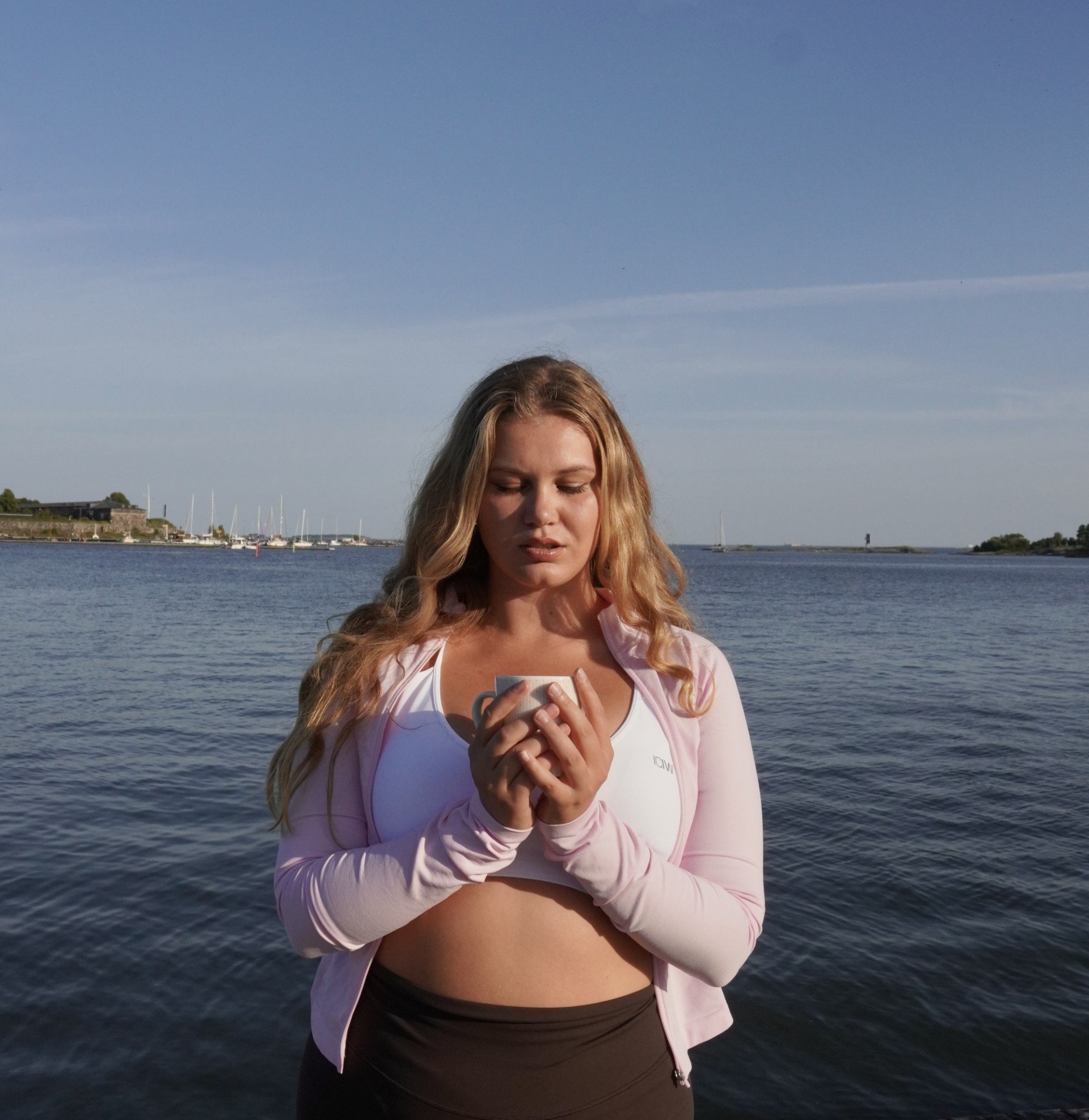 A young woman with long, wavy blonde hair looking at her smartphone while standing outdoors near water with boats in the background.