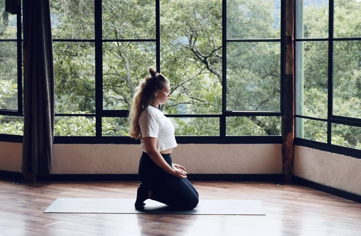 A woman practicing yin yoga kneeling on a yoga mat in front of large windows with a view of green trees.