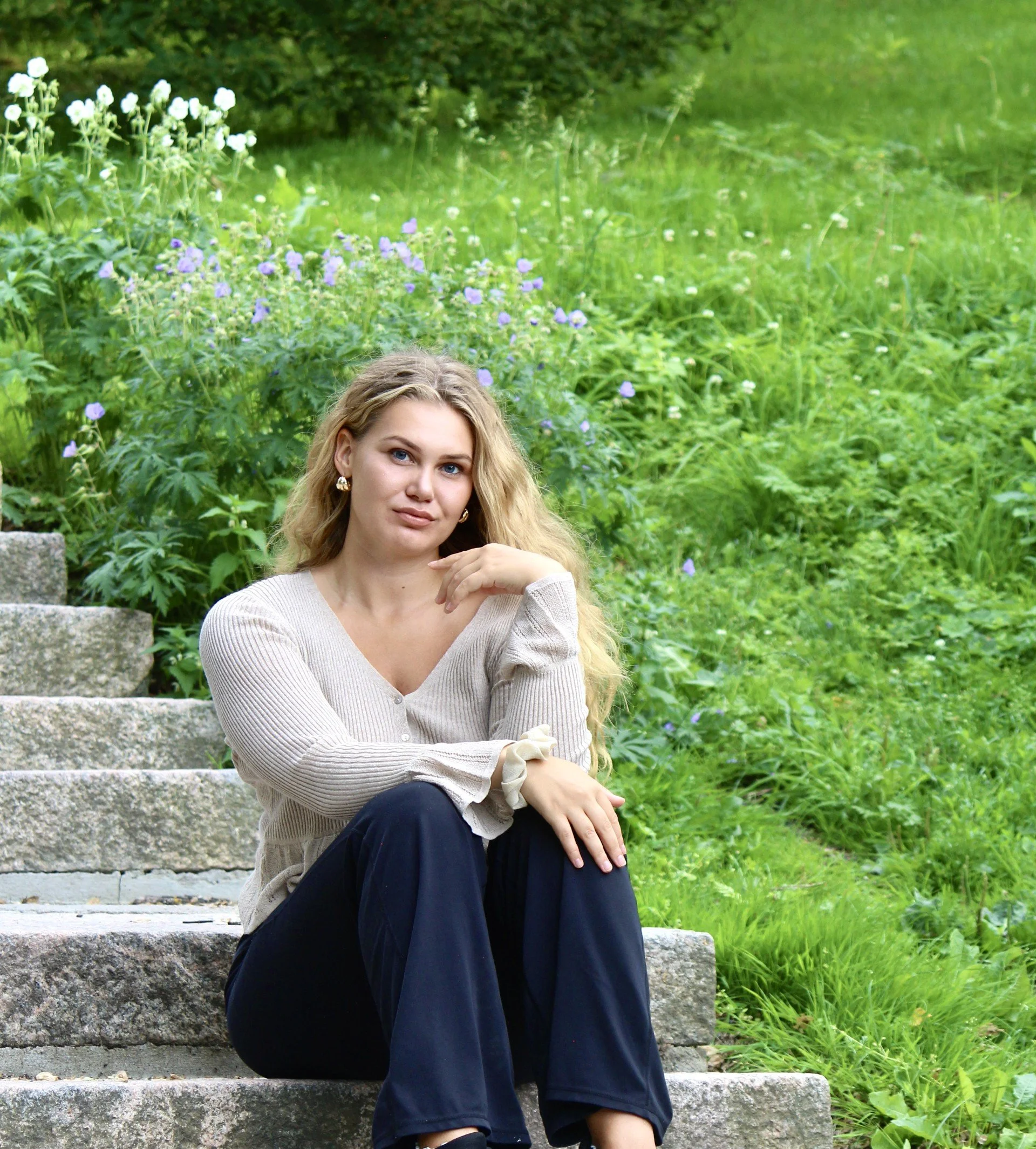 A woman with long curly blonde hair and blue eyes sitting on stone steps outdoors, surrounded by green grass and plants, wearing a cream-colored sweater, dark pants, and gold earrings.