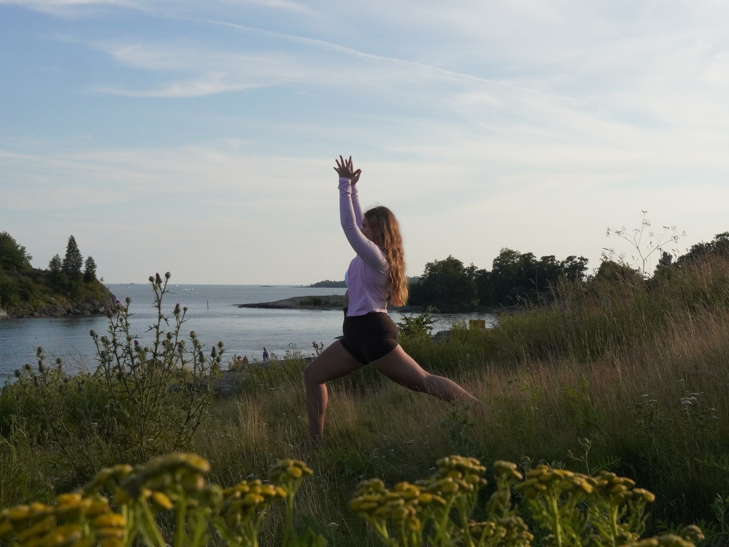 A woman in a yoga pose outdoors on a grassy area near a body of water with trees and a cloudy sky in the background.