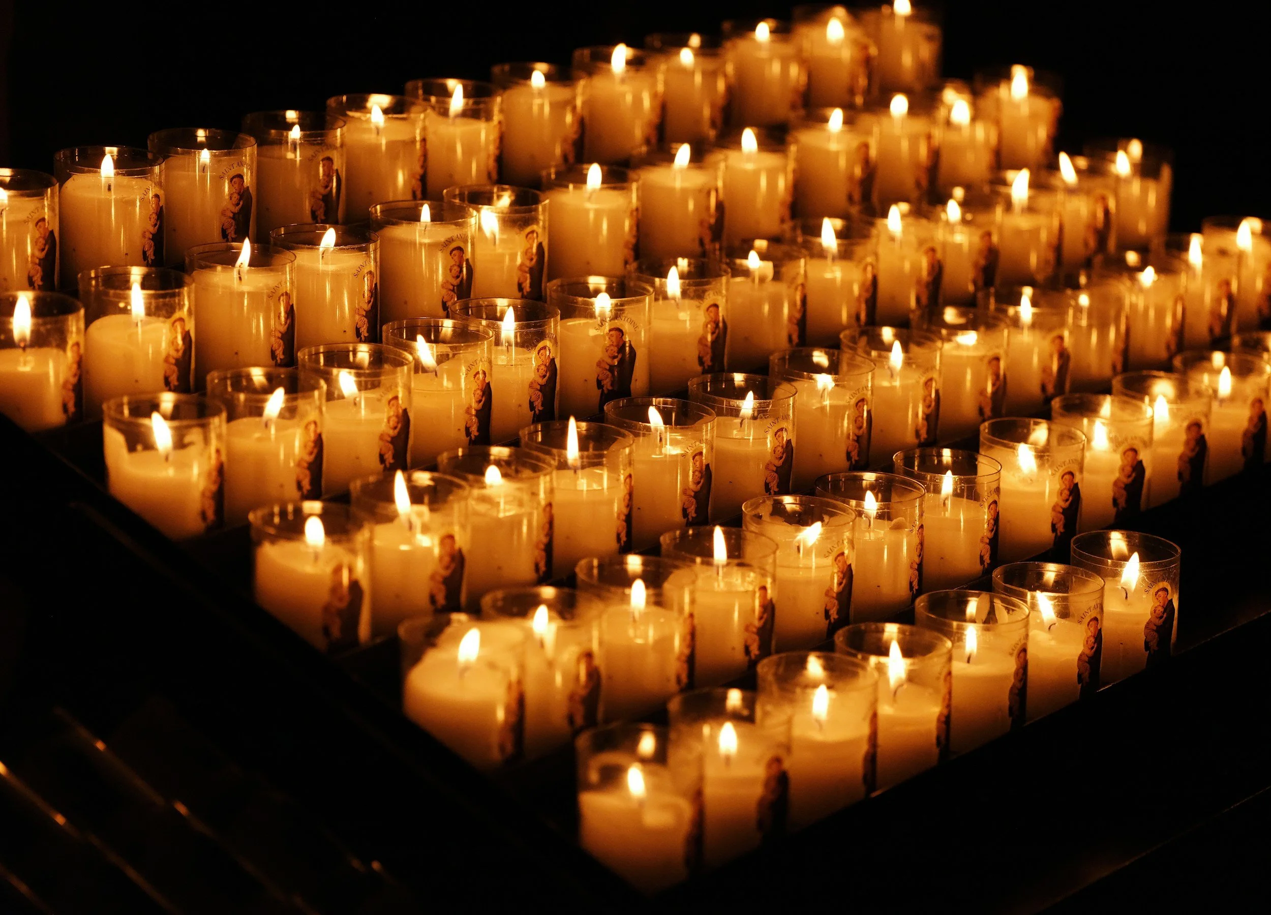 Numerous lit candles with religious images on them arranged in a grid, likely used for a memorial or religious ceremony.