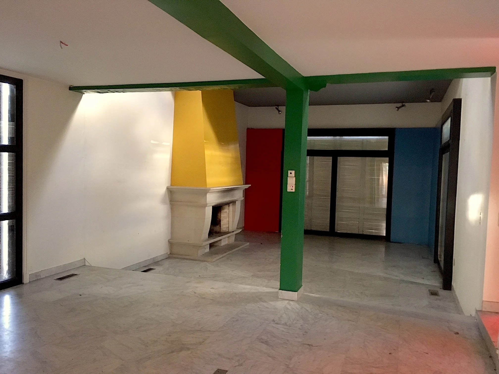 Intérieur d'une maison avec cheminée en pierre, murs colorés en jaune, rouge, bleu, et structure en vert, sol en marbre, fenêtres et porte en verre.