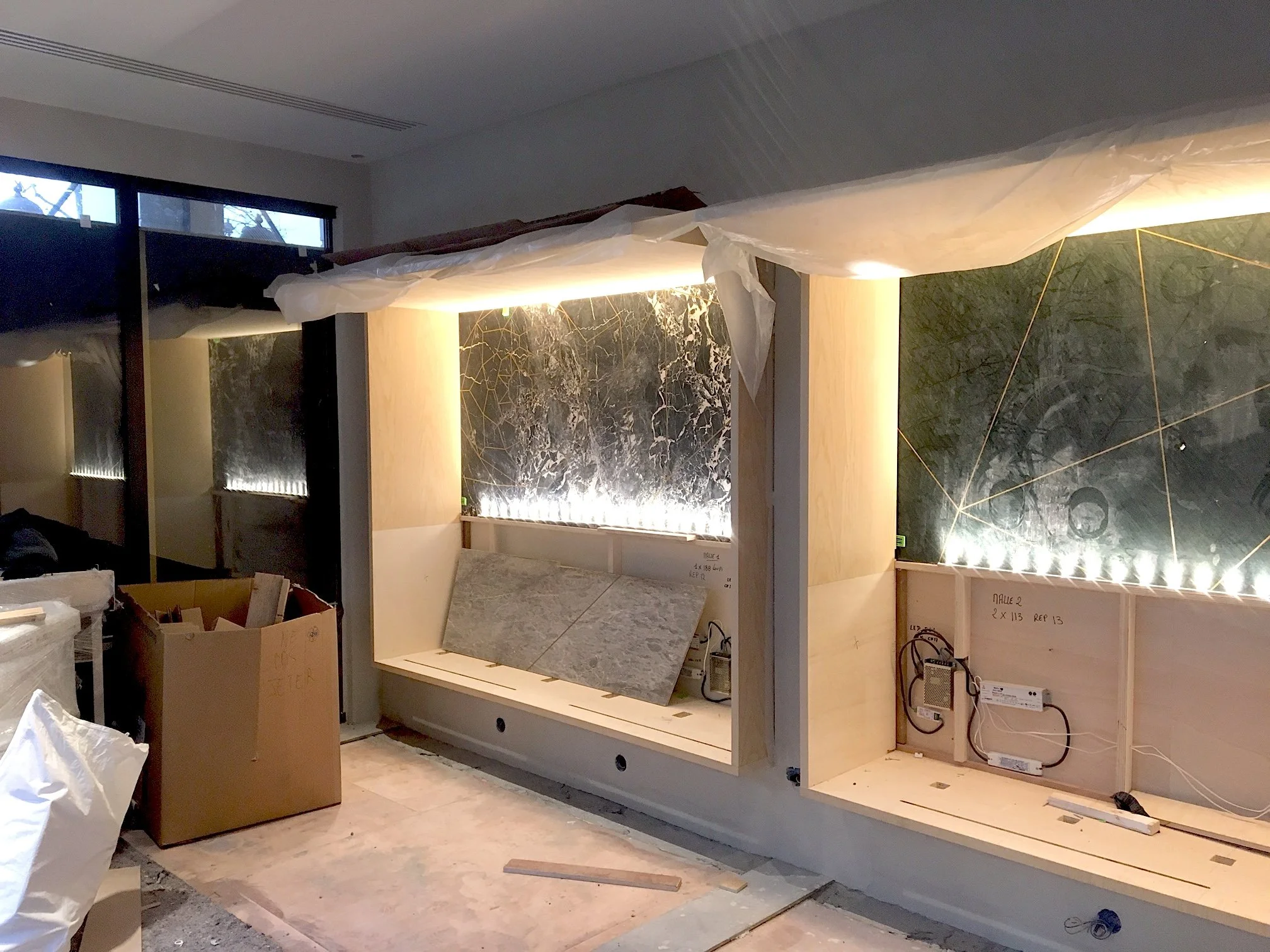 Salle en construction avec murs en marbre, éclairage intégré, et structure en bois pour meubles ou installations, avec des matériaux et outils de construction visibles.