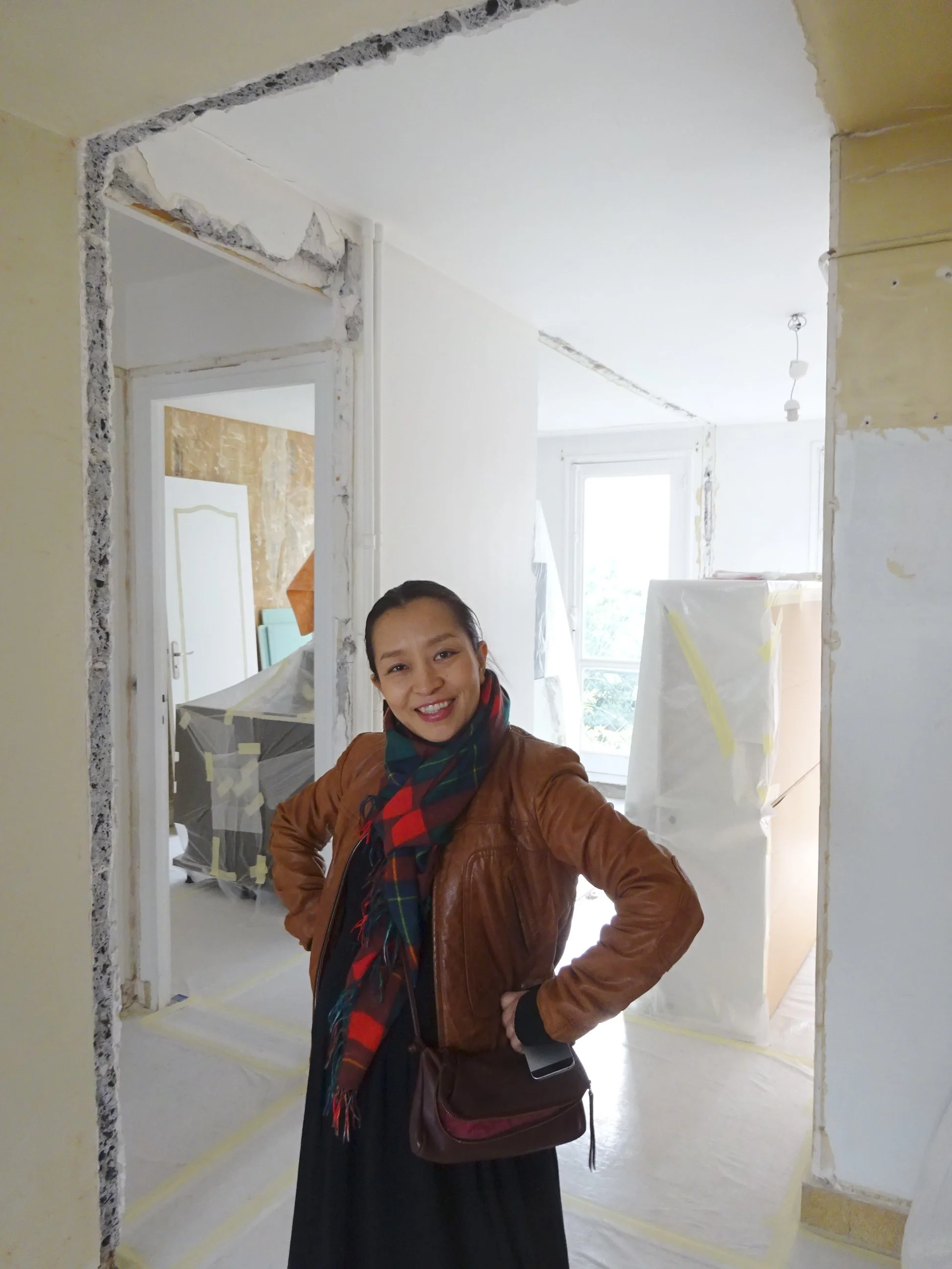 Une femme souriante portant un manteau en cuir marron et une écharpe à carreaux, posant dans une pièce en cours de rénovation.