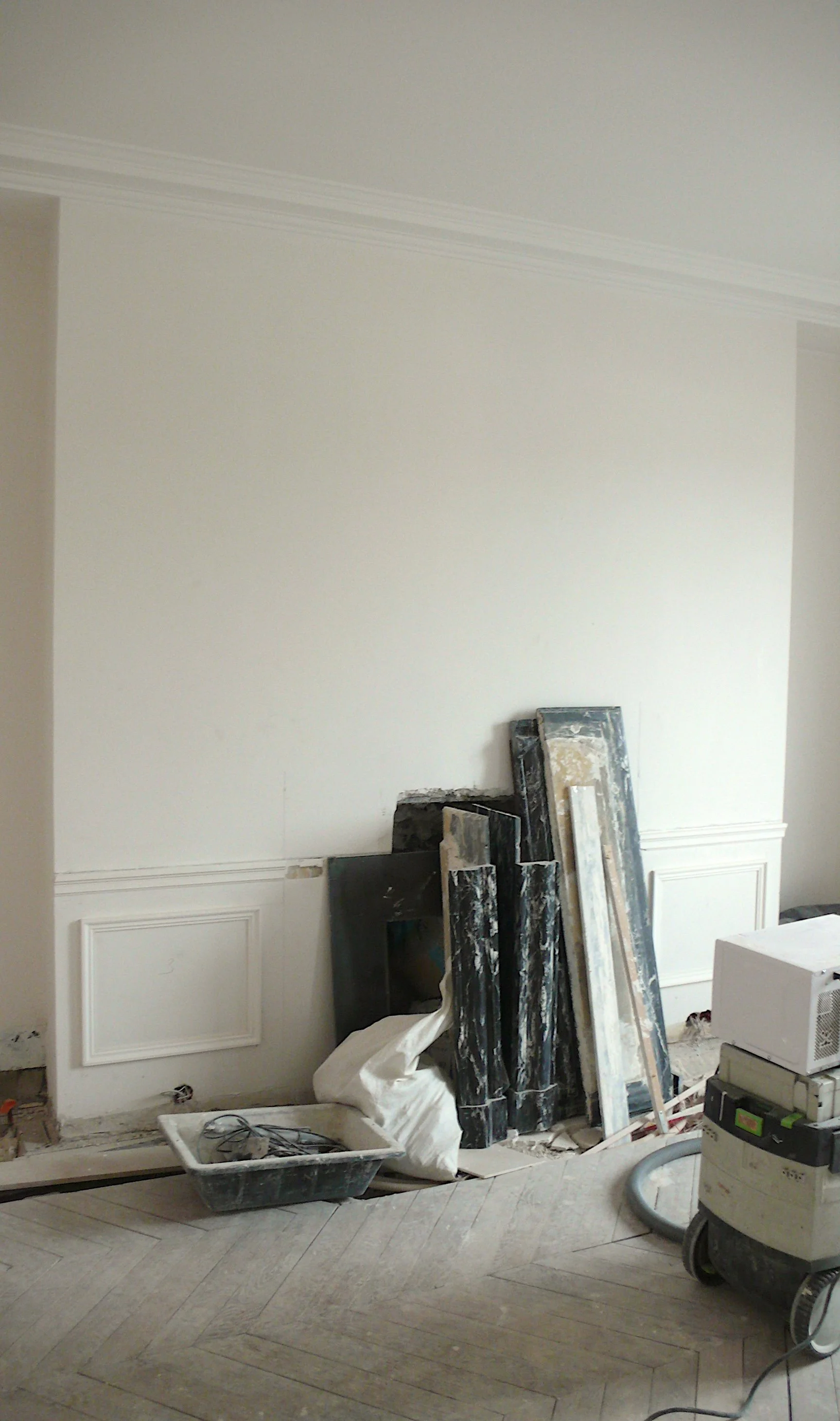 Chambre en rénovation avec des plaques de marbre empilées contre le mur blanc, un seau, des câbles, une boîte à outils et un ventilateur de chantier