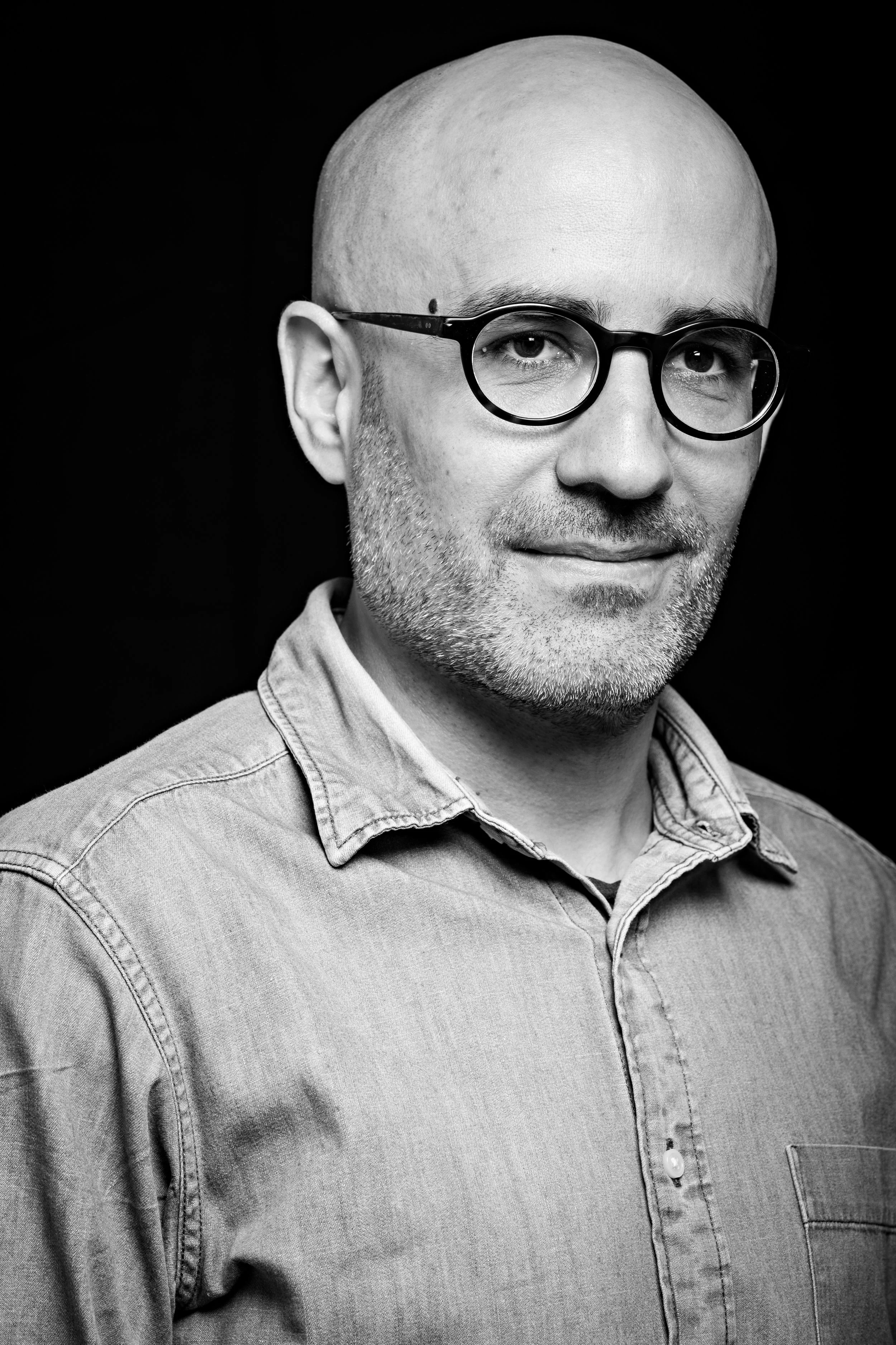 Portrait en noir et blanc d'un homme portant des lunettes rondes, une barbe courte, une tête rasée et un chemisier en denim avec un fond noir.